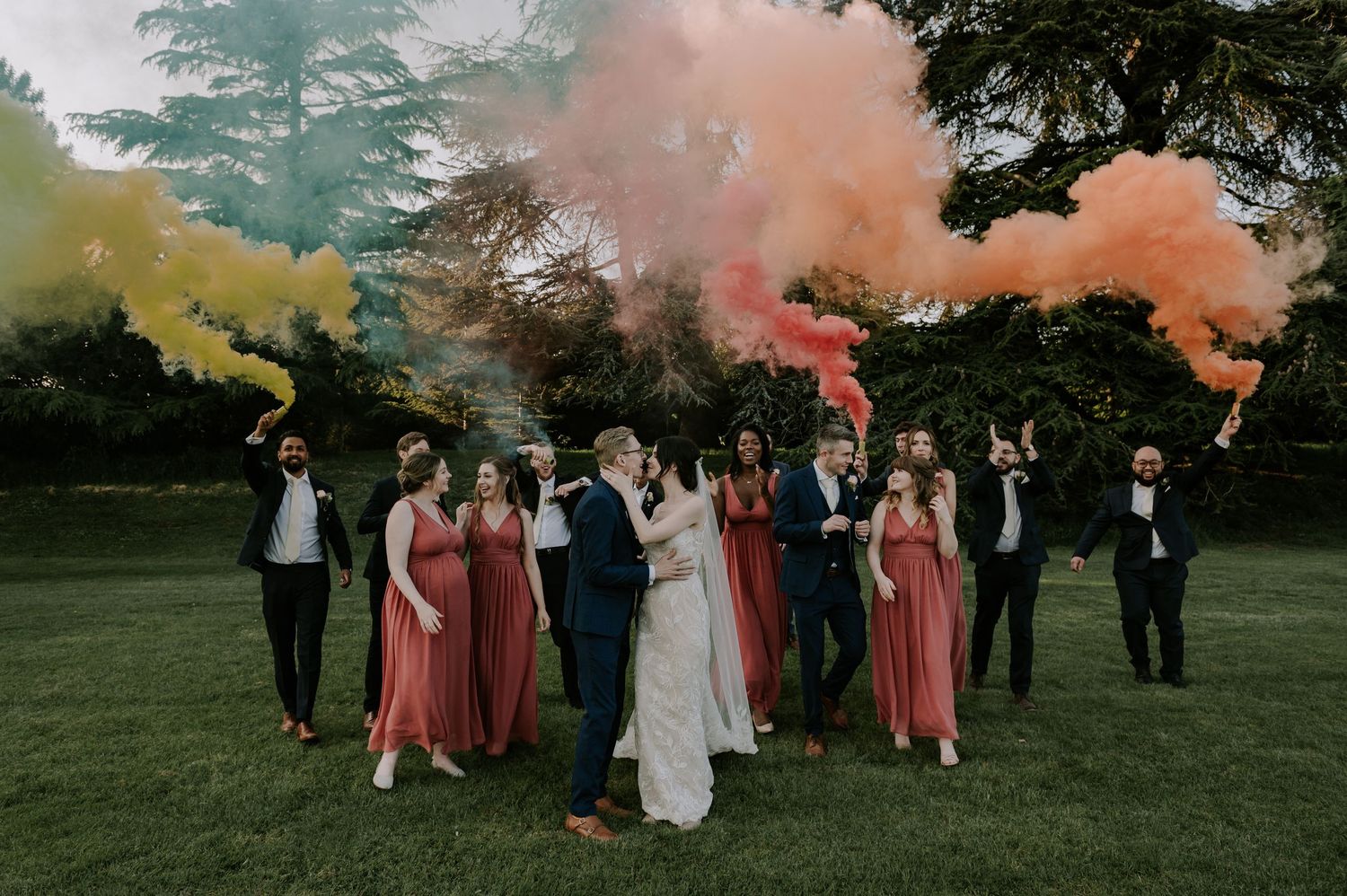 Wedding party stands on green lawn holding colorful smoke bombs creating orange, pink and yellow clouds against trees.