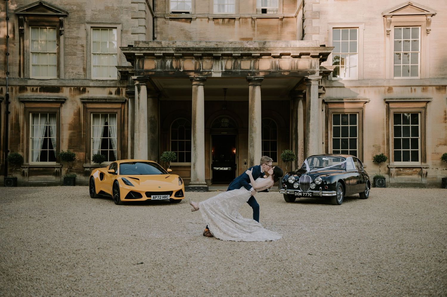 A couple dances in front of a historic mansion with a yellow McLaren and vintage car parked on the gravel driveway.