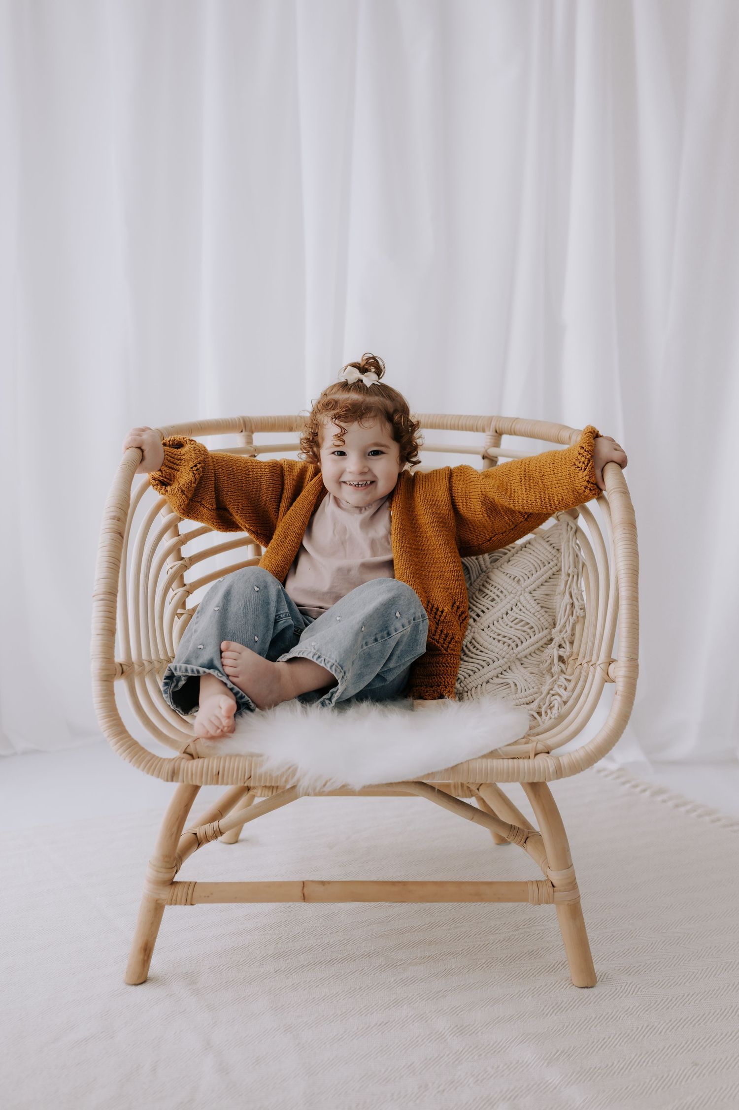 Child in mustard cardigan sitting in rattan chair with sheepskin, studio, Melton Mowbray.