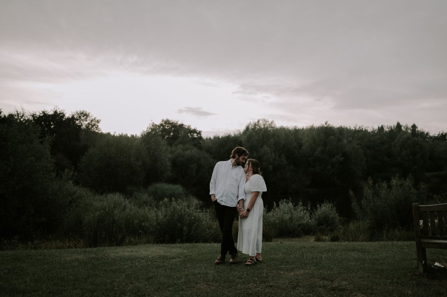 A couple stands together on a grassy field at dusk with dark trees silhouetted in the background.