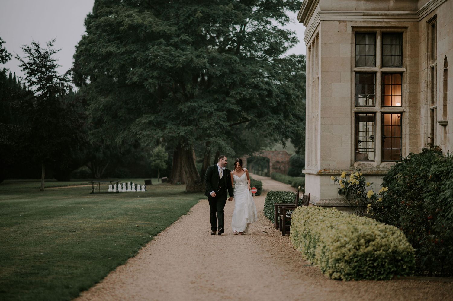 Newlywed couple walks along a scenic garden path outside an elegant stone mansion during their wedding celebration.
