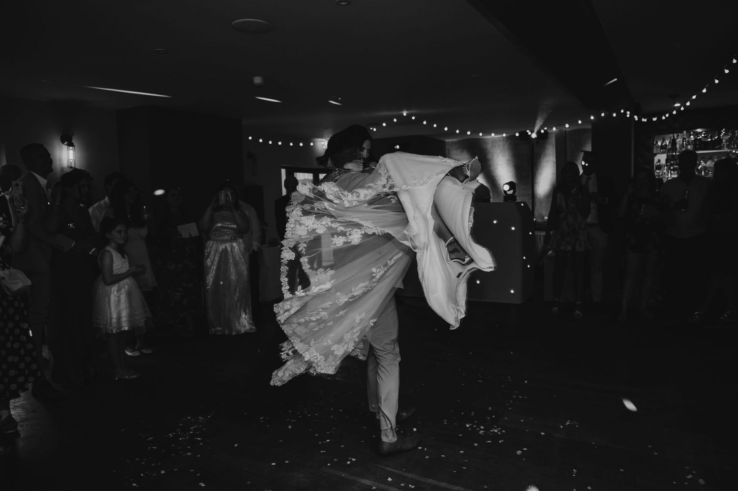 Groom lifting and twirling bride on dance floor beneath string lights, lace dress in black and white – Chequers Inn, Grantham wedding.