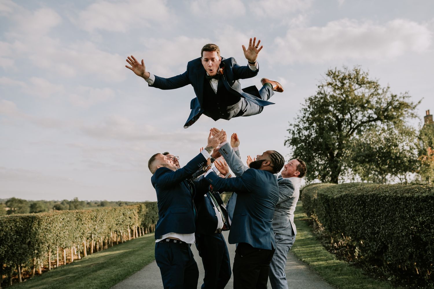 Groomsmen toss a groom in the air against a blue sky during a fun wedding photo session.