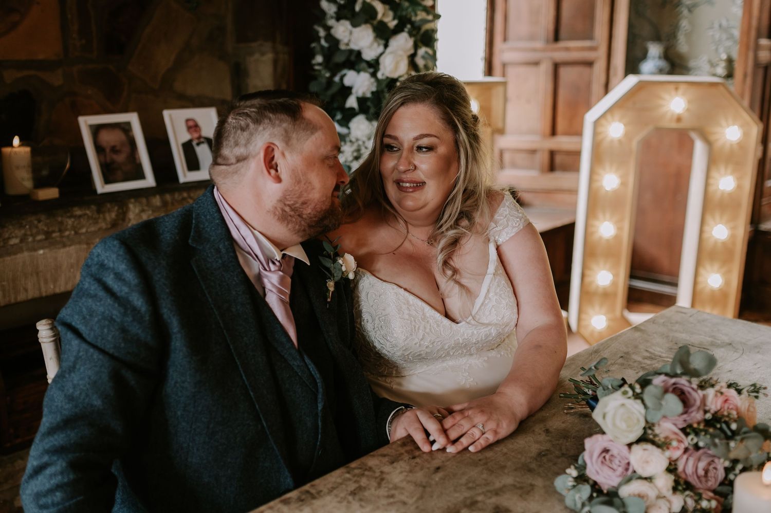 An intimate wedding moment captured between newlyweds seated at a table with pink floral decorations and lit marquee lights.