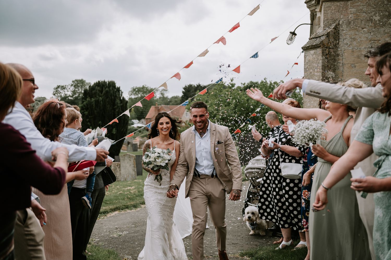 Wedding guests shower confetti over a happy couple exiting their outdoor countryside ceremony.