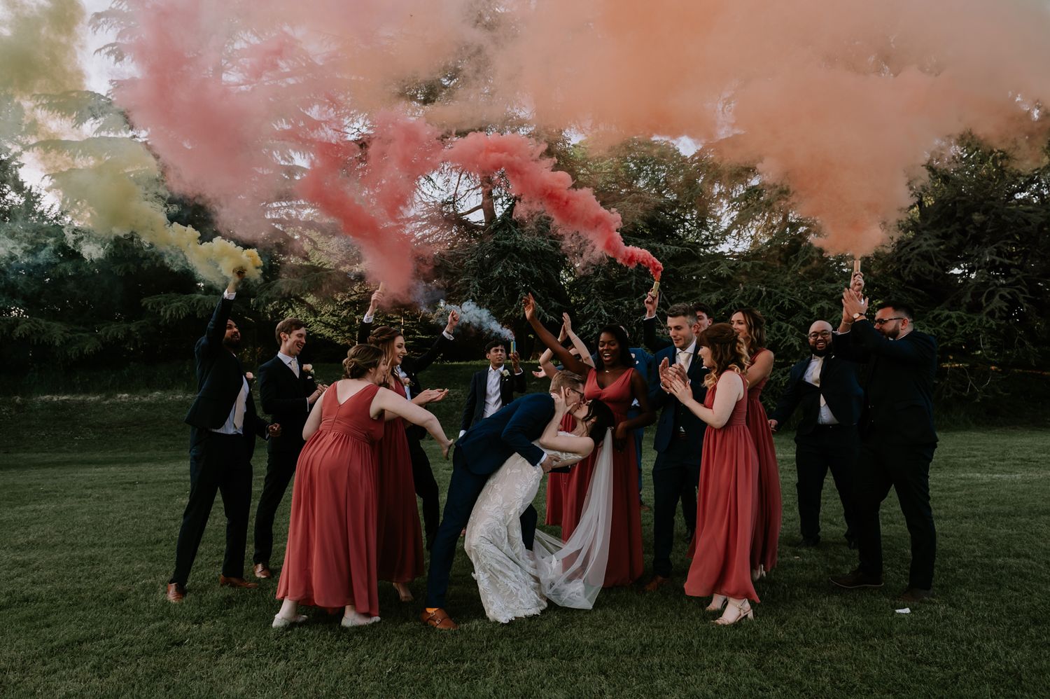 Wedding party celebrates on green lawn with pink smoke bombs creating a dramatic sunset atmosphere.