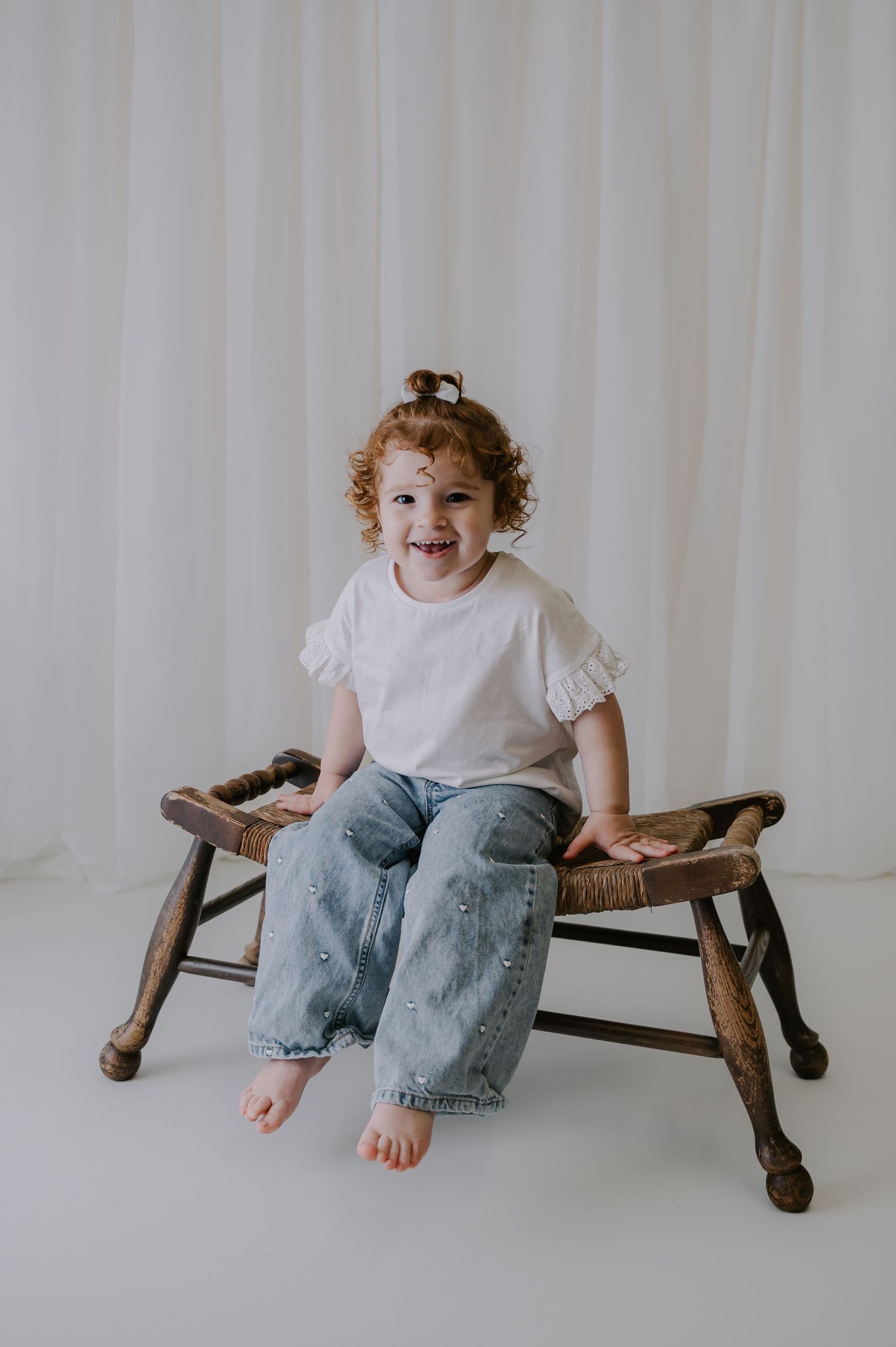 Child in white shirt and jeans sitting on wooden bench against white backdrop in minimalist studio setting.