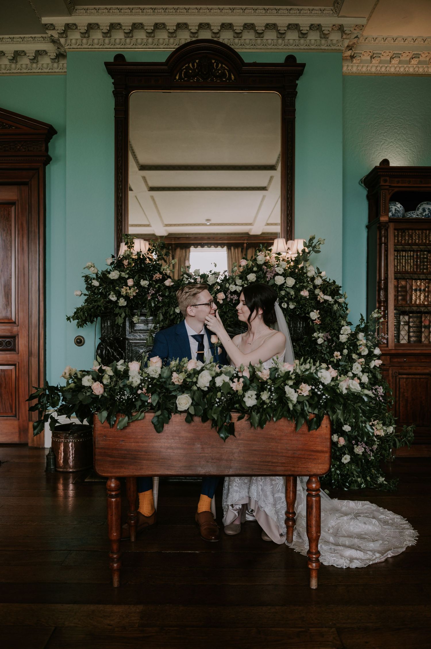A bridal gown elegantly draped over dark wooden furniture in an ornate room with turquoise walls and floral arrangements.