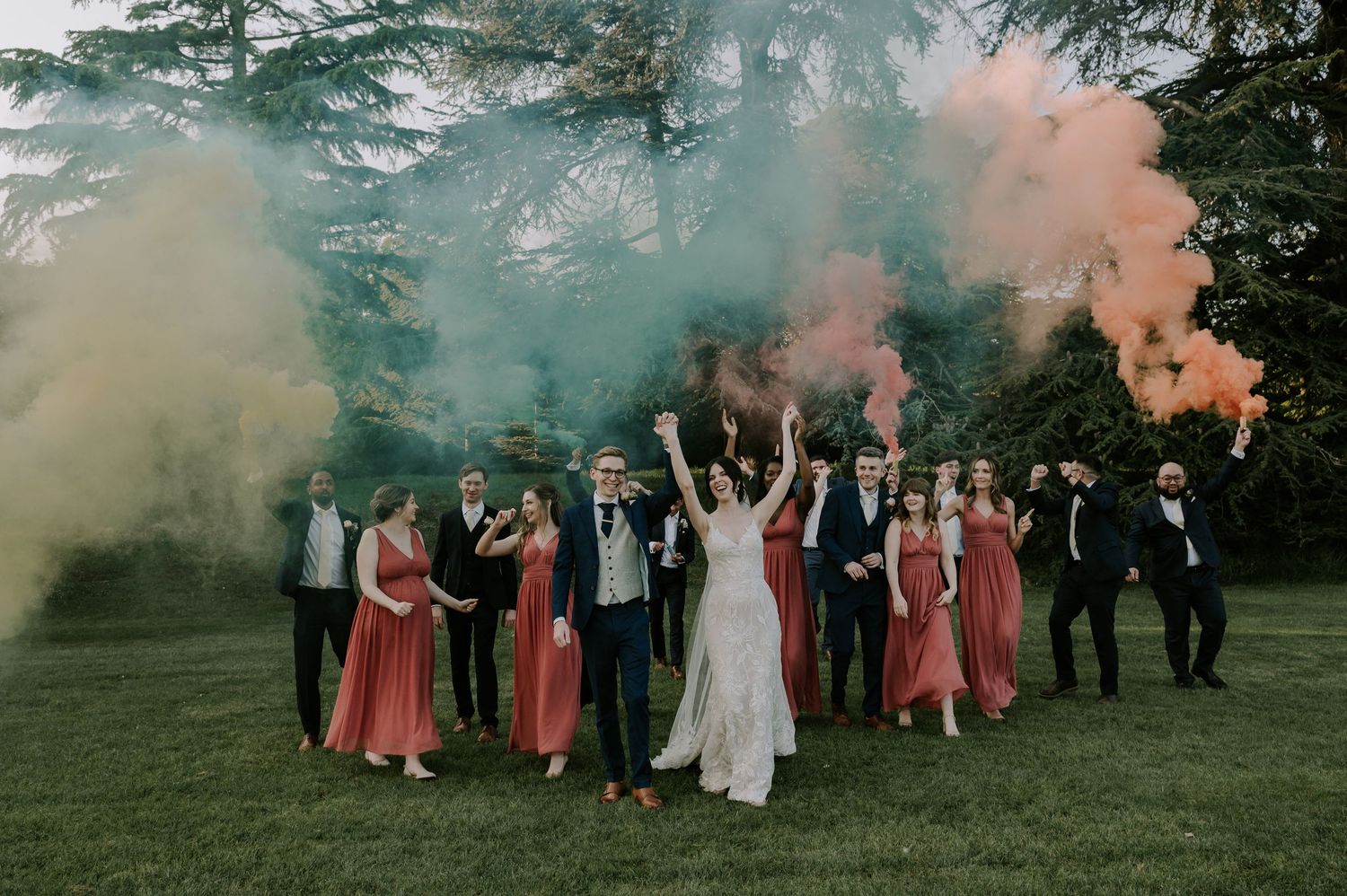 A wedding party stands together outdoors with colored smoke bombs creating a dreamy pink and green atmosphere at sunset.