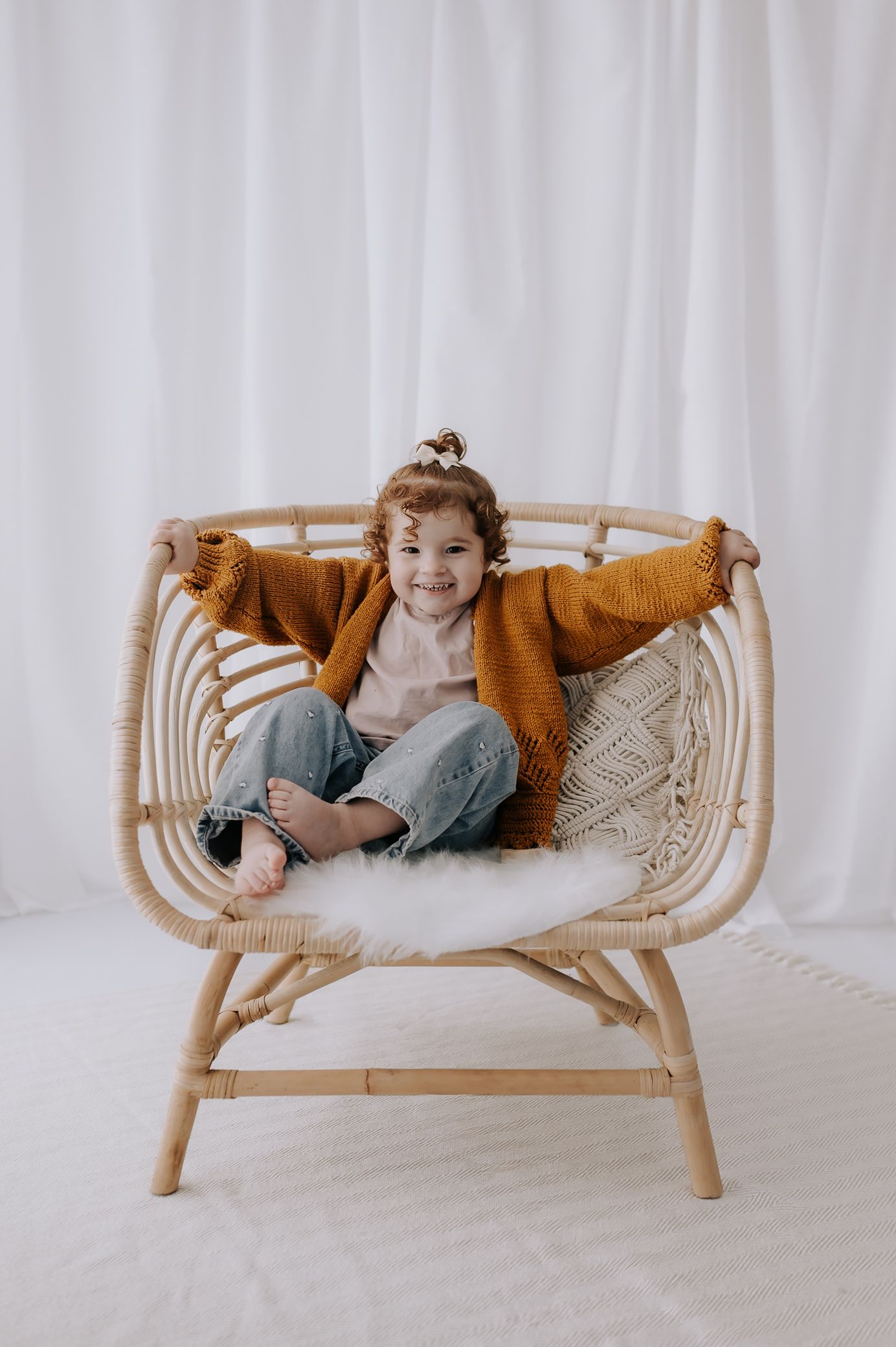 Child in mustard cardigan and jeans sitting happily in rattan chair with white fur cushion in studio – Melton Mowbray children’s photoshoot