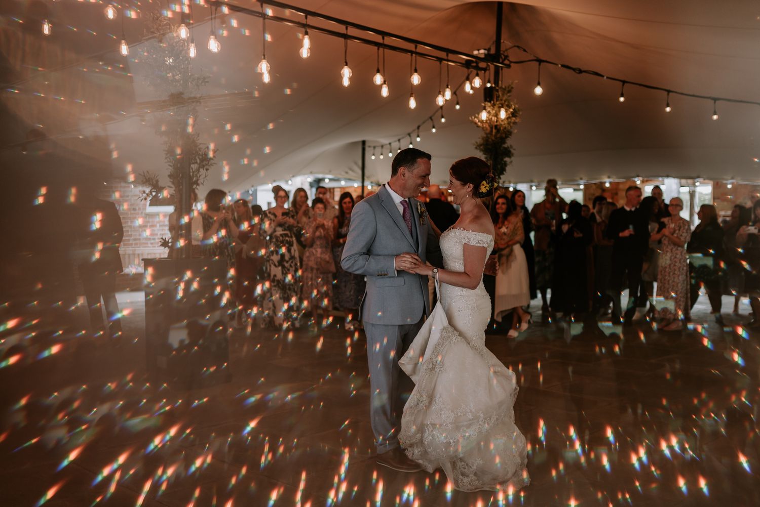 A couple shares a romantic first dance under twinkling lights in a wedding reception tent as guests look on.