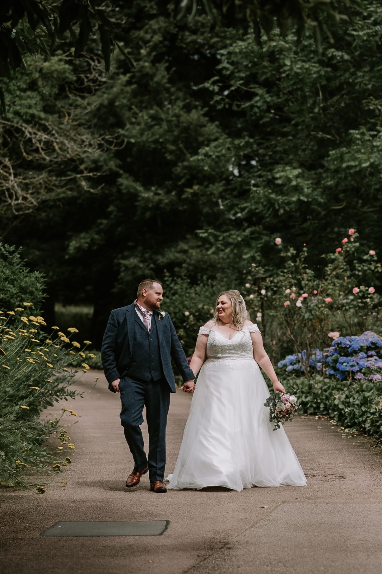 A couple walks hand in hand on a garden path surrounded by lush greenery and blooming flowers on their wedding day.