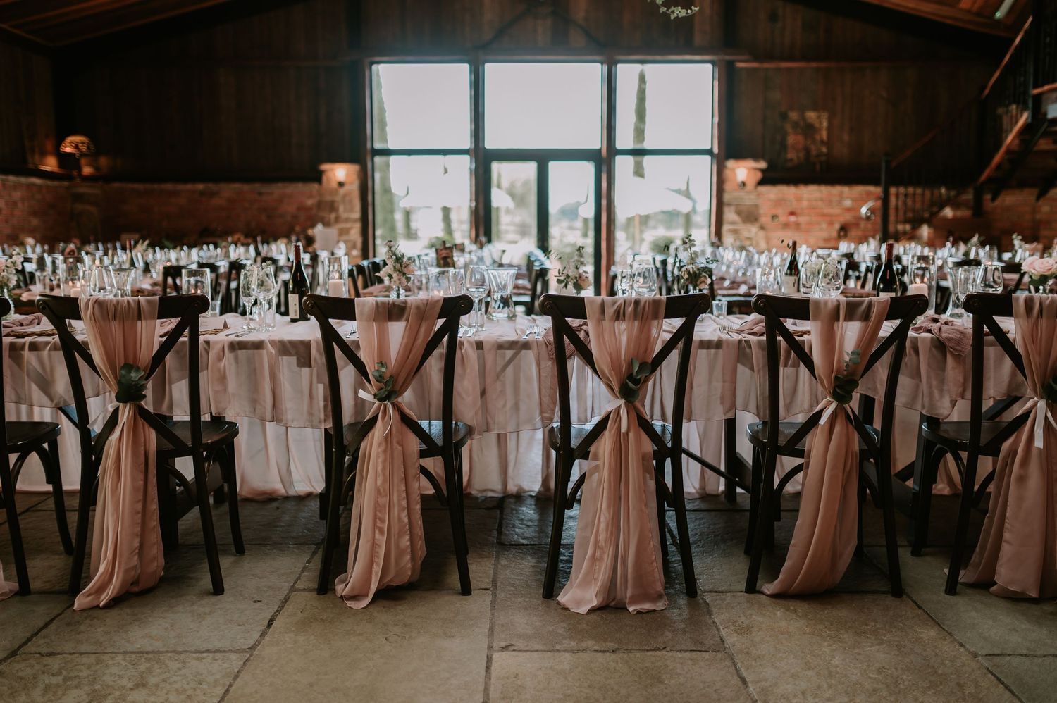 Rustic wedding reception tables with rose gold draping tablecloths and black cross-back chairs in an industrial venue.