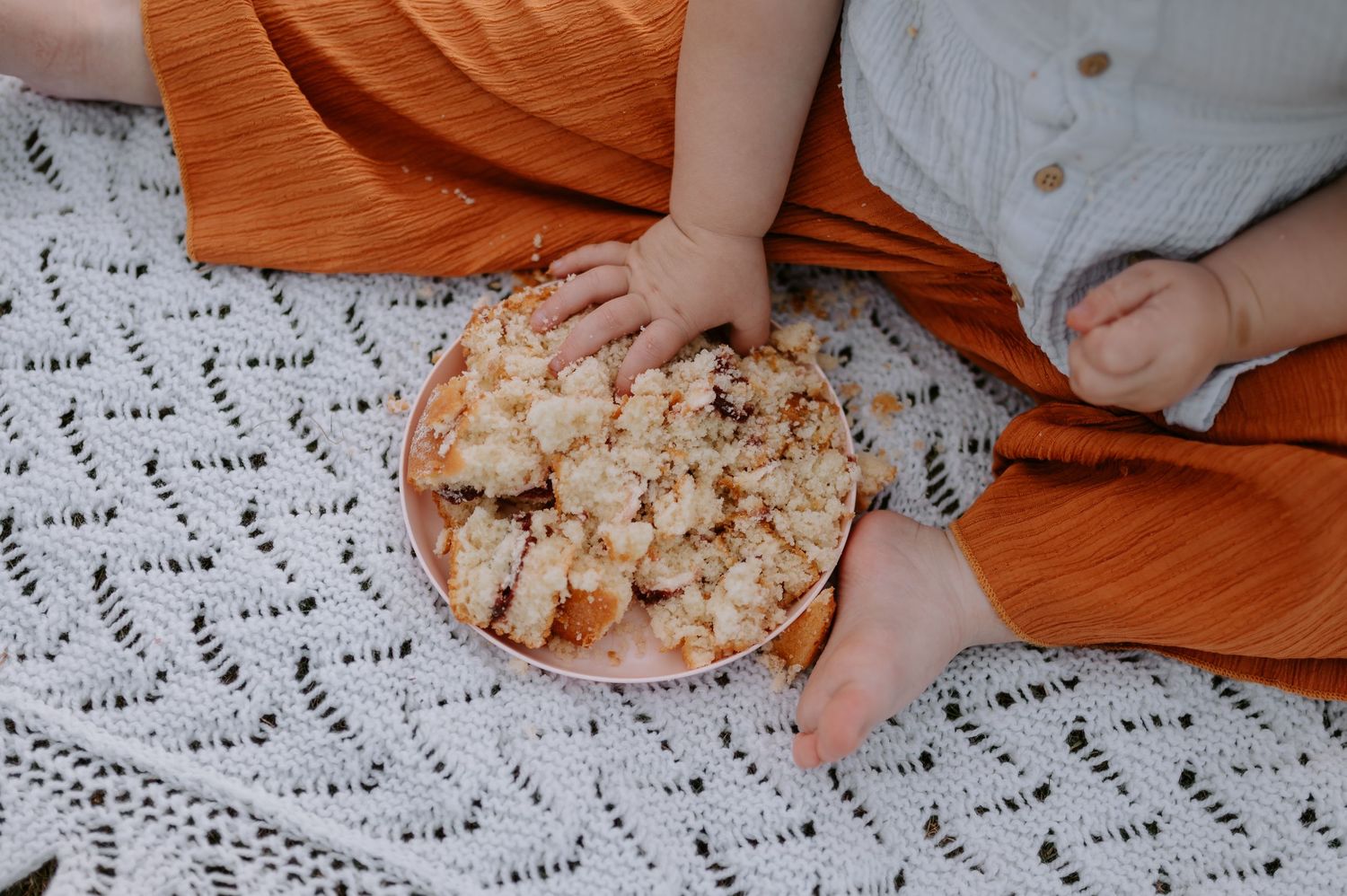 A crumbled cake spread on a pink plate between the legs of orange trousers during a cake smash photoshoot