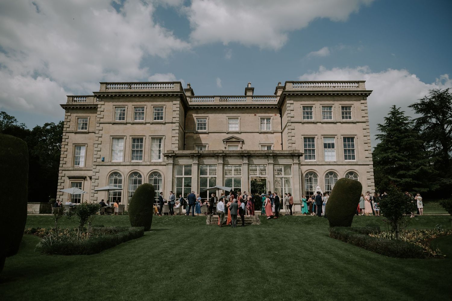 Elegant historic manor house with neoclassical architecture set on manicured lawns under cloudy blue skies.