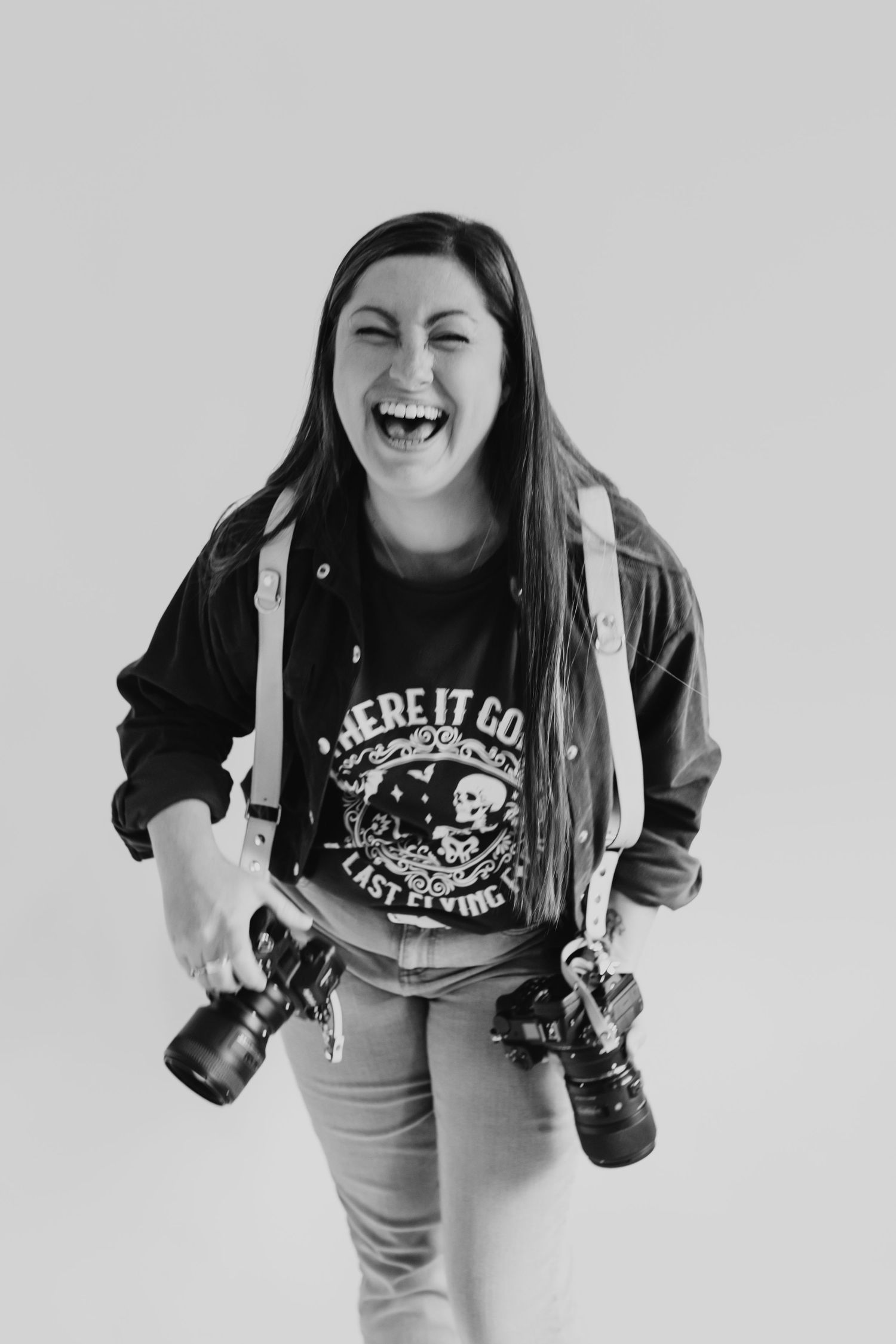 Black and white portrait of photographer laughing while holding two DSLR cameras against plain background.