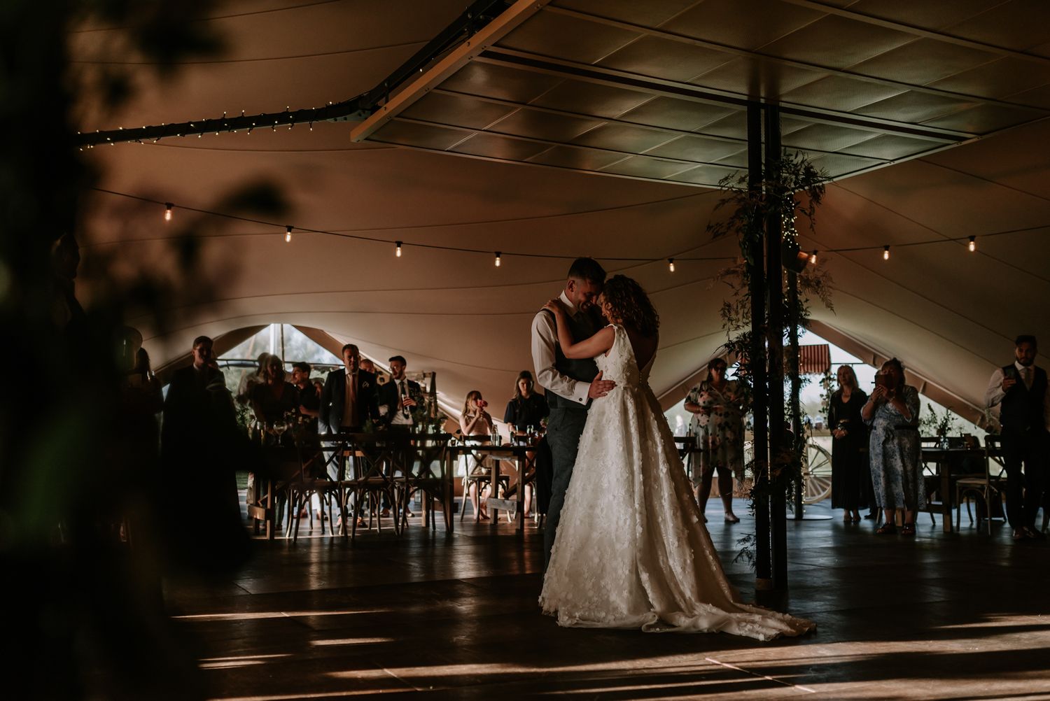 A couple embraces during their first dance under a romantic tent reception with string lights and elegant table settings.