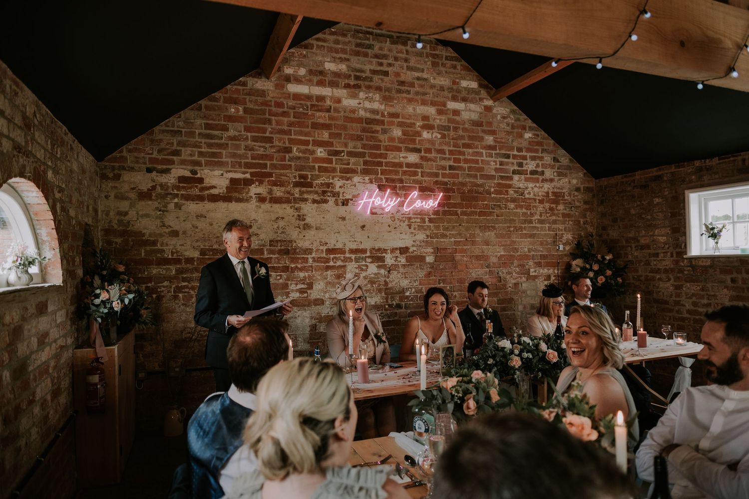Wedding reception guests gather in a rustic brick venue with a pink neon sign glowing on the wall during speeches.
