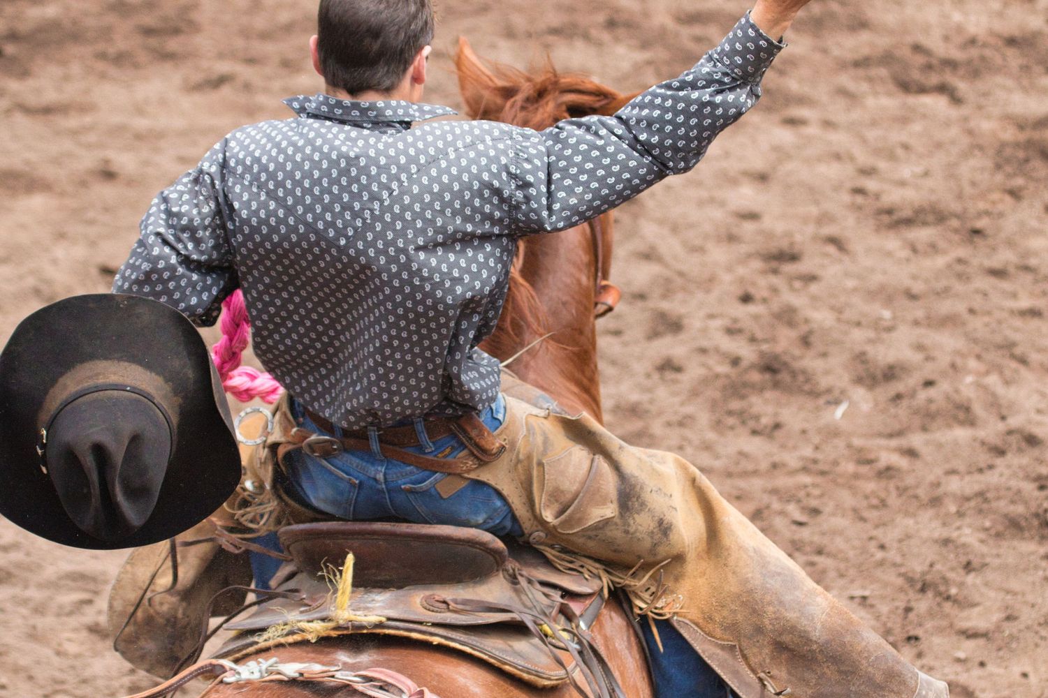 A rodeo cowboy rides a bucking horse in a patterned shirt, hat flying, during a competitive rodeo event.