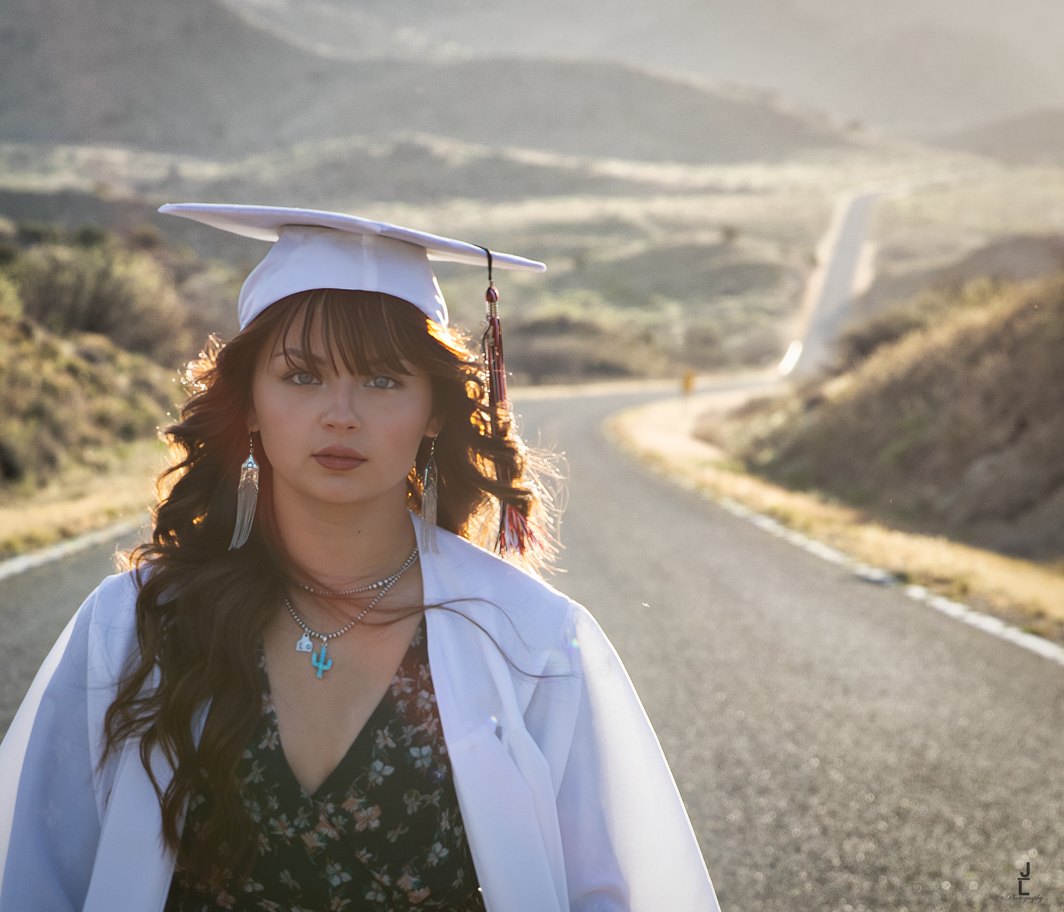 Graduate in white cap and gown walks along a scenic winding desert highway at golden hour.
