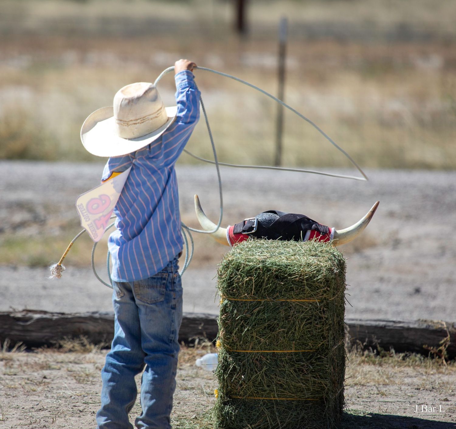 A young cowboy practices lasso roping on a hay bale with fake bull horns outdoors.