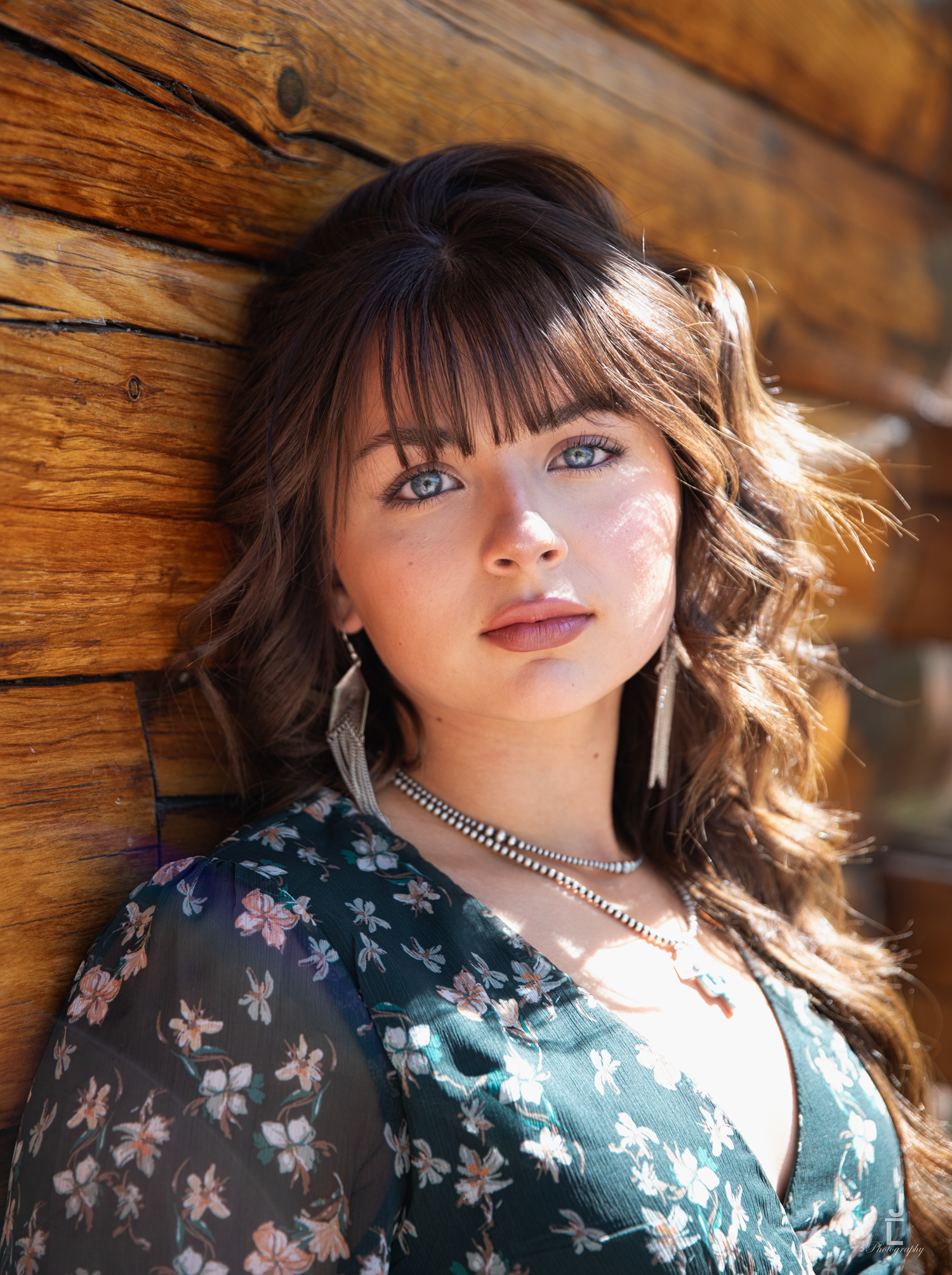 Young woman with blue eyes and wavy hair leans against rustic wooden log cabin wall.