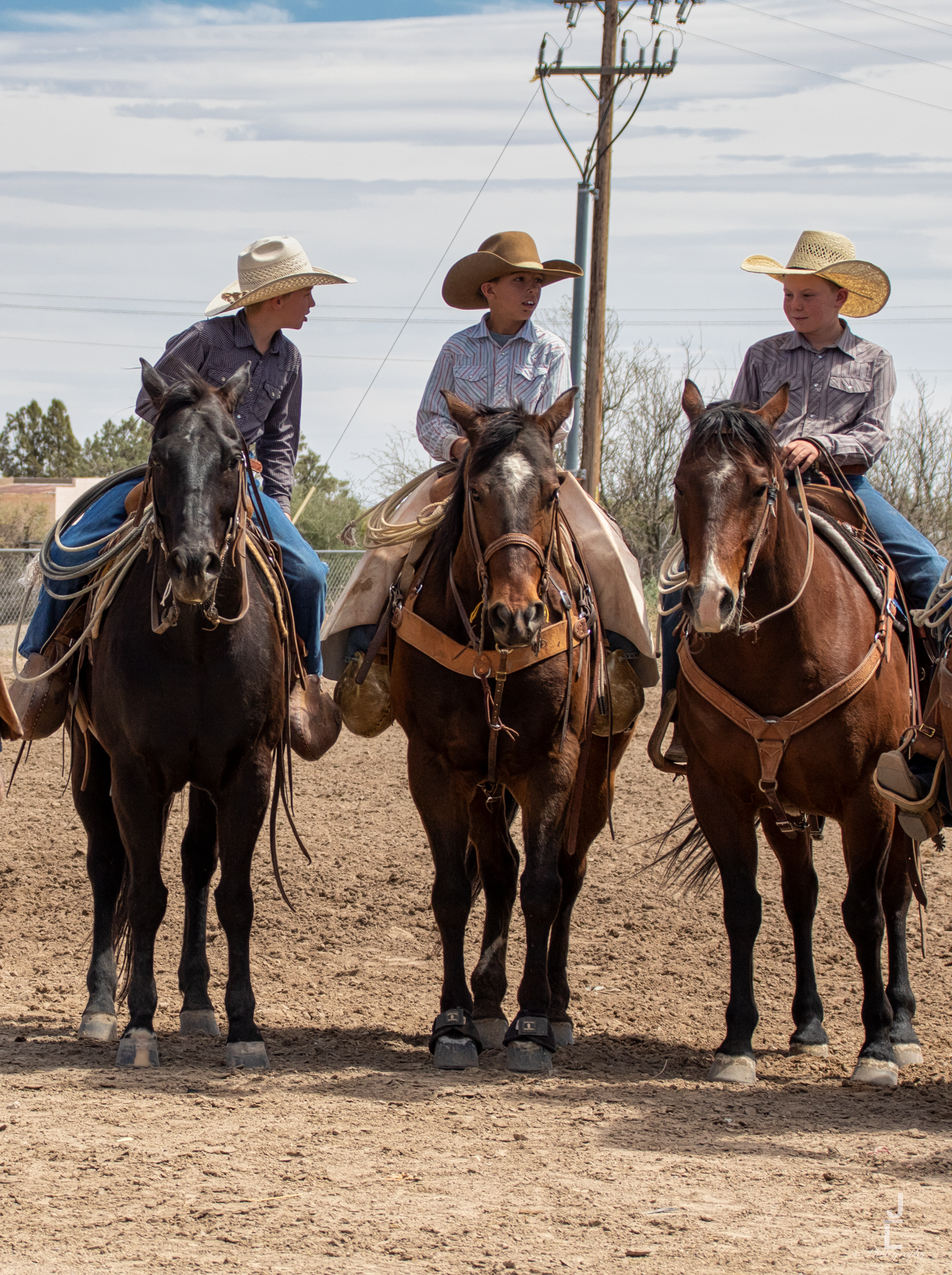 Three cowboys wearing cowboy hats sit on horseback side by side in a dirt arena.