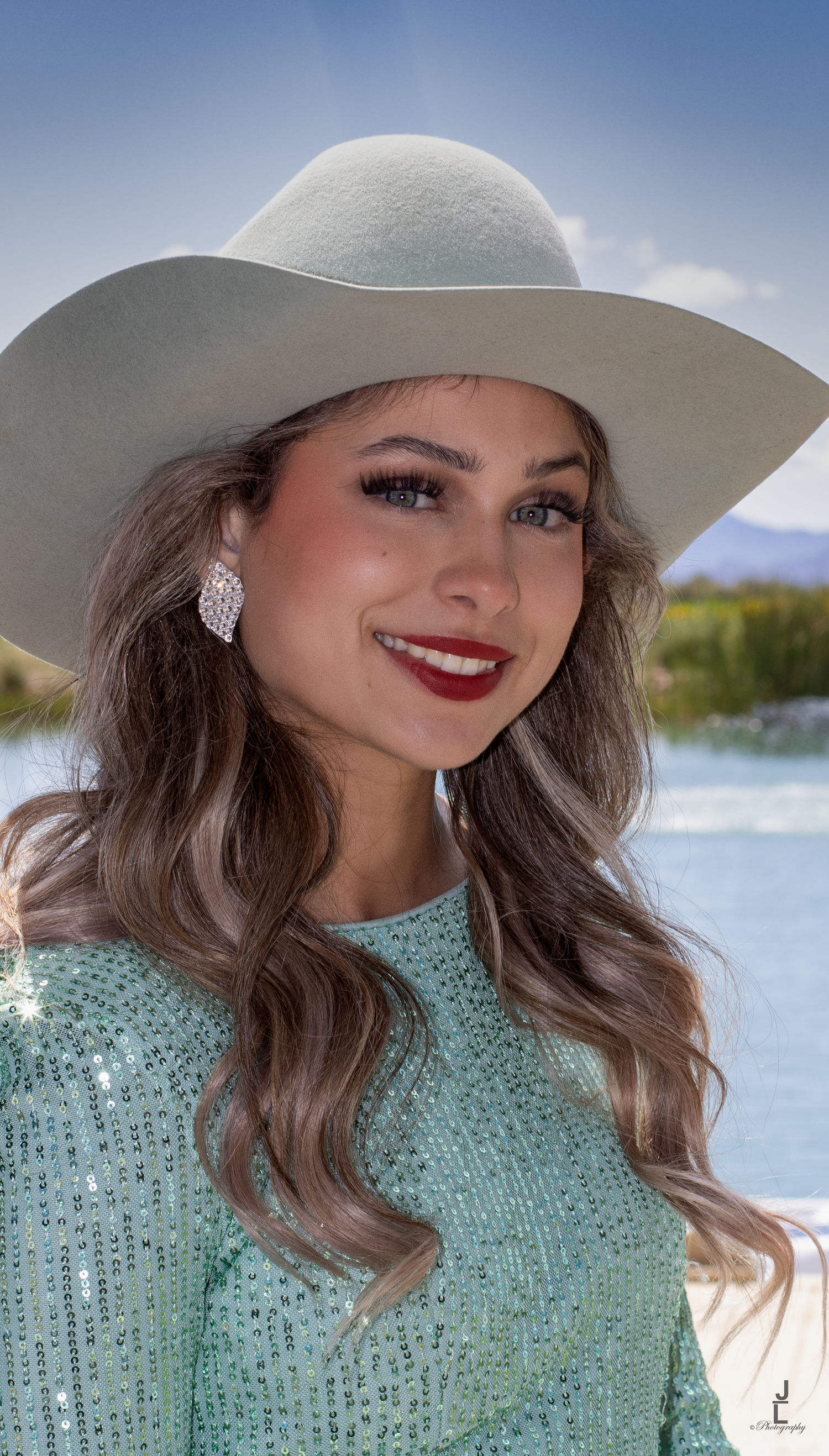 Smiling young woman wearing a white cowboy hat and mint sequin top with chandelier earrings outdoors.