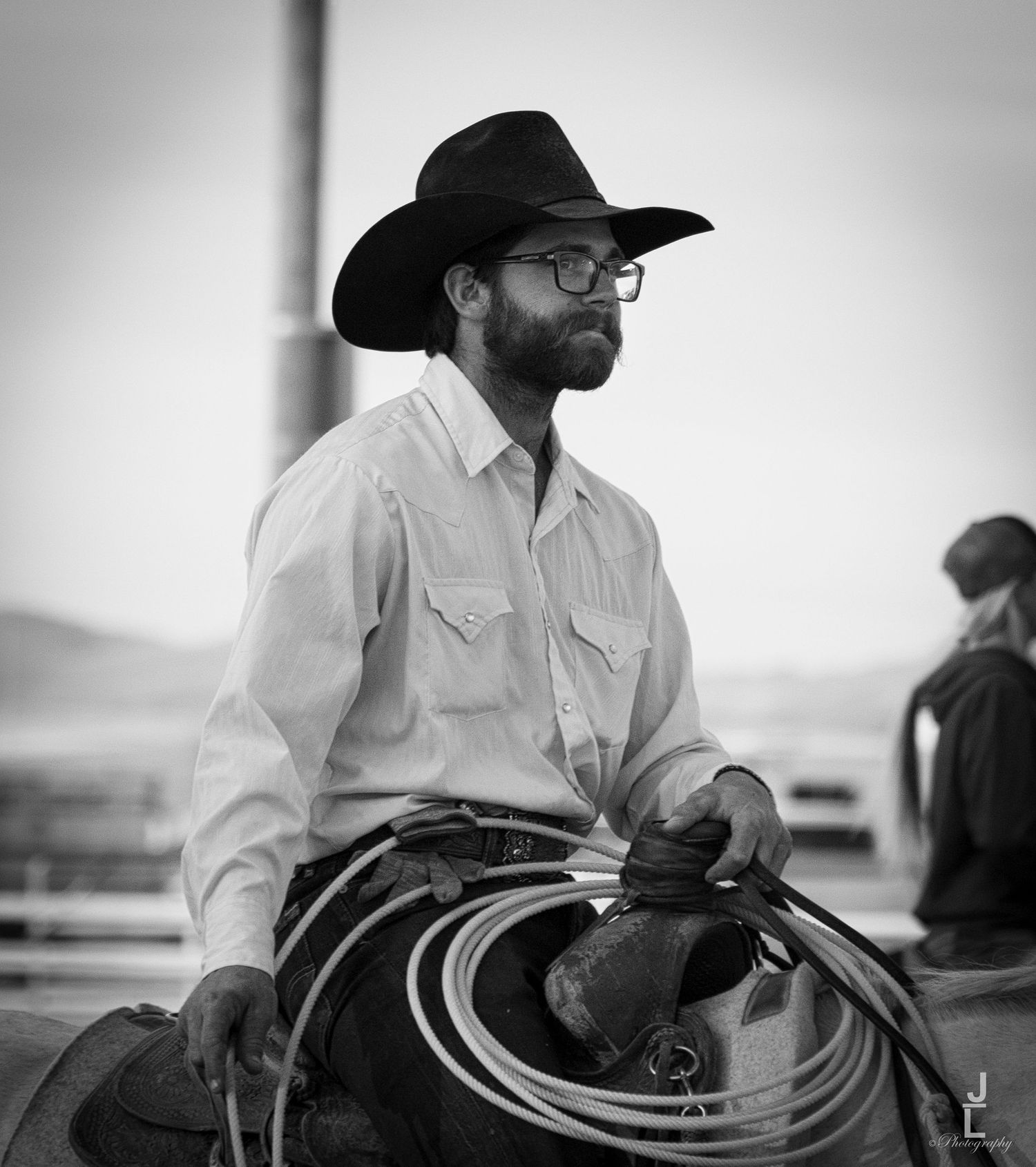 Black and white portrait of a bearded cowboy in black hat holding a lasso rope on horseback.