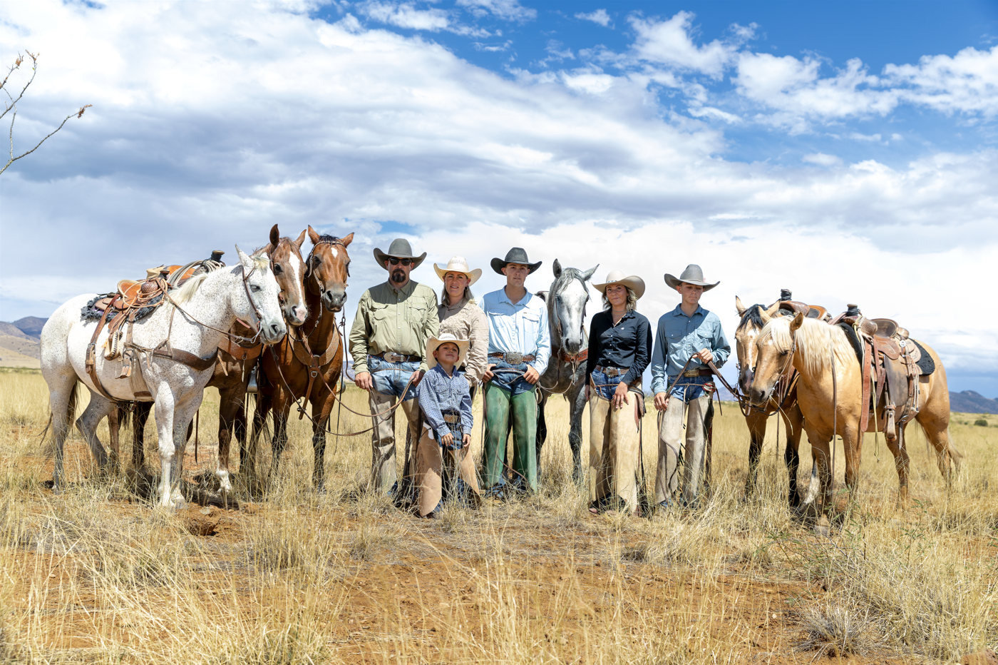 Group of cowboys and cowgirls with horses pose in golden grassland under cloudy sky.