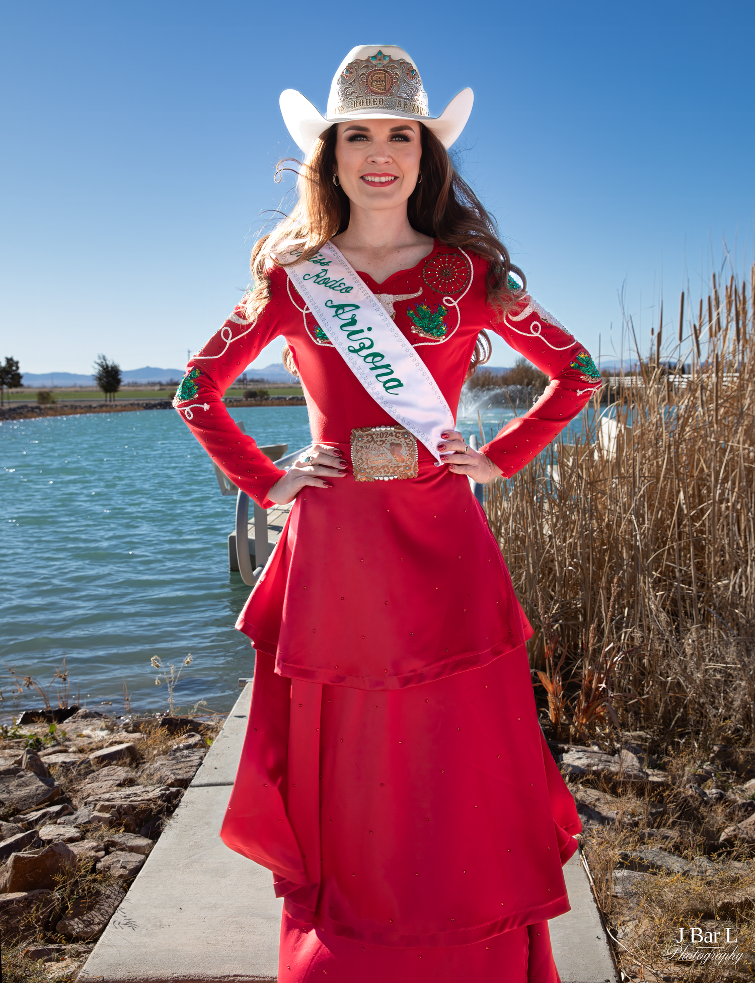Miss Arizona rodeo queen poses in red western outfit with white cowboy hat near a lake.