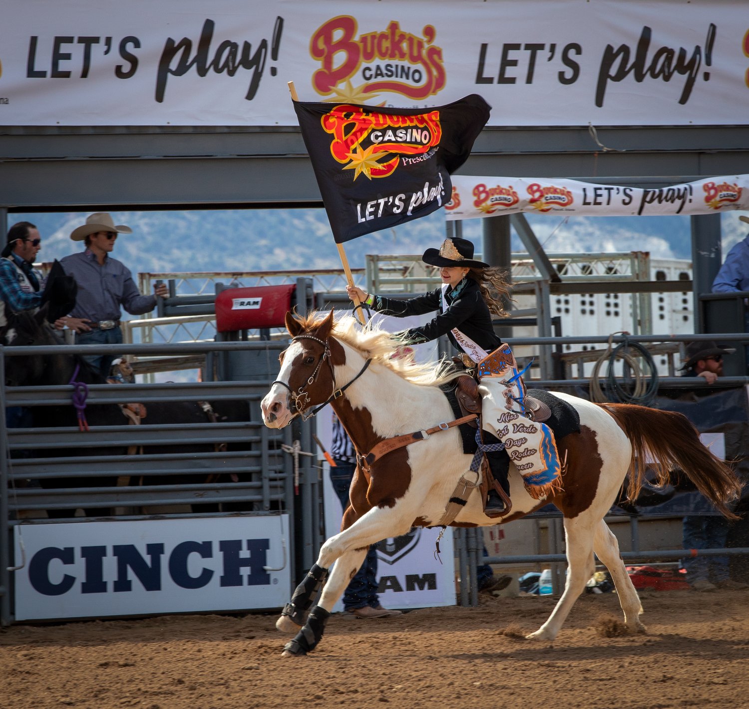 Rodeo queen rides a paint horse carrying a Bucky's Casino flag at an indoor rodeo arena.