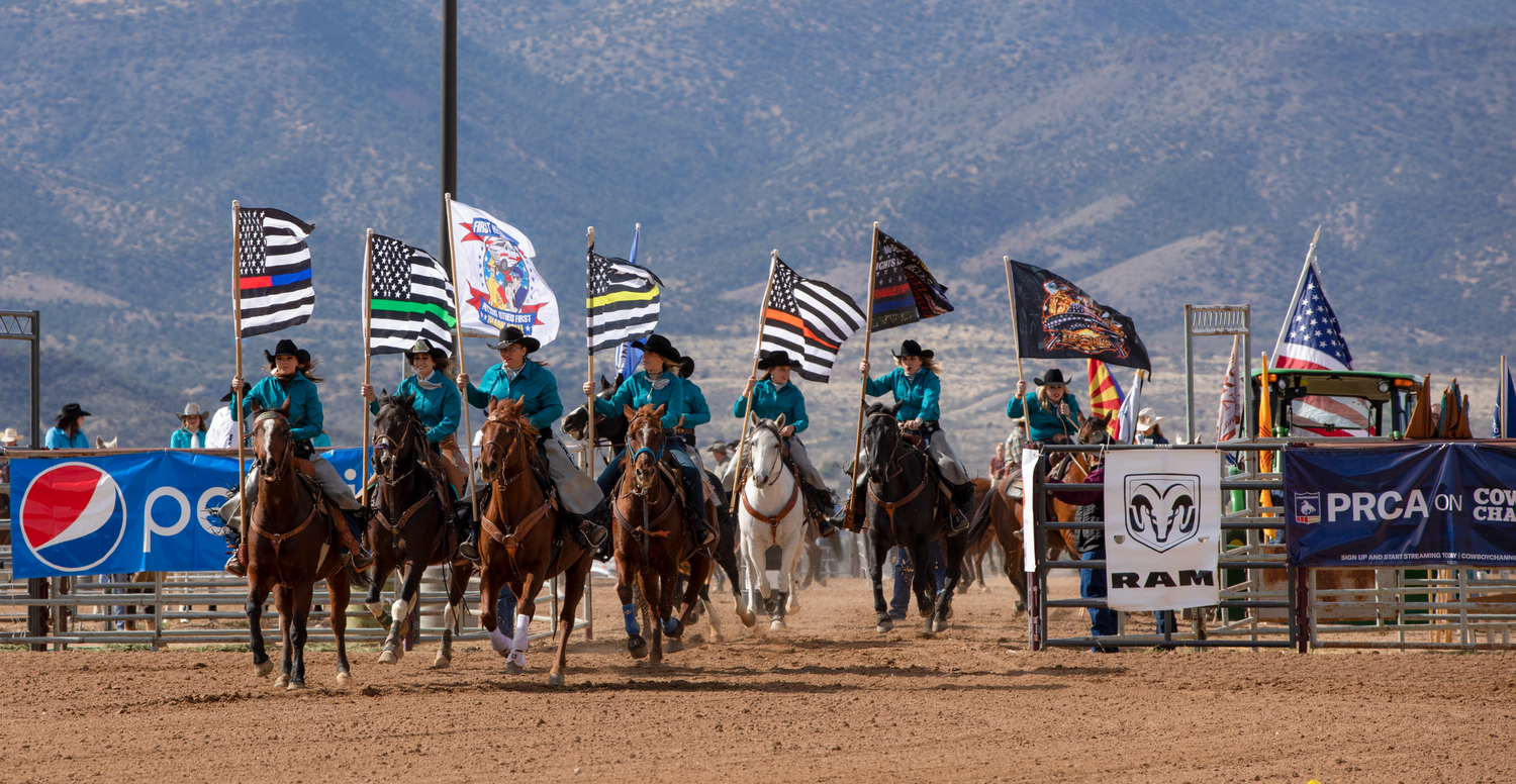 Riders on horseback carrying colorful flags gallop through a PRCA rodeo arena with mountains behind.