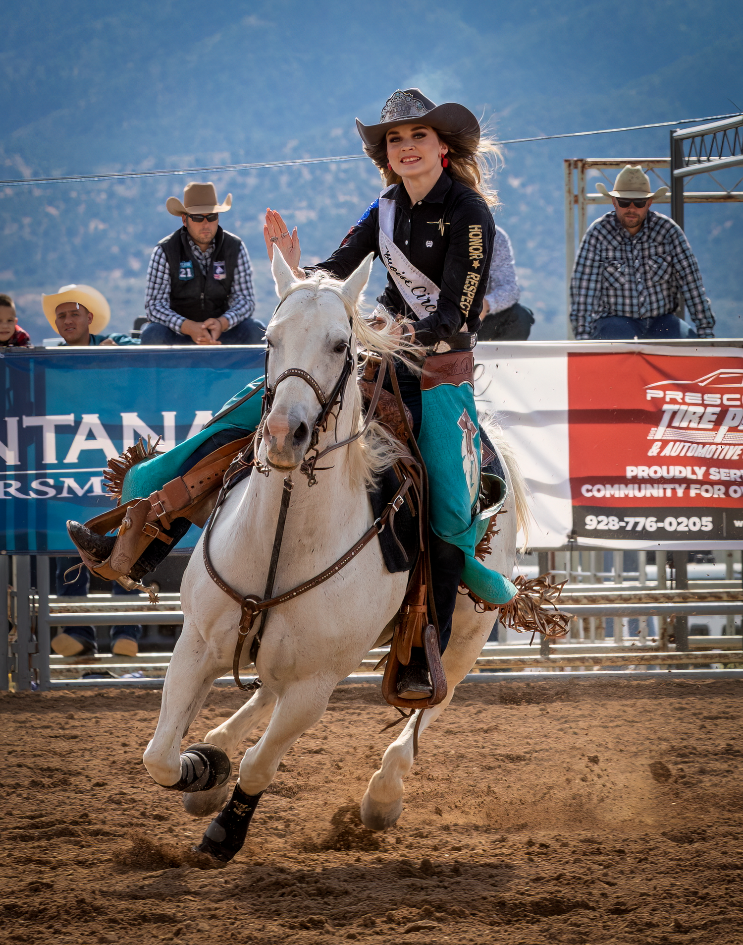 Rodeo queen in teal outfit and cowboy hat rides white horse at full gallop in a dirt arena.