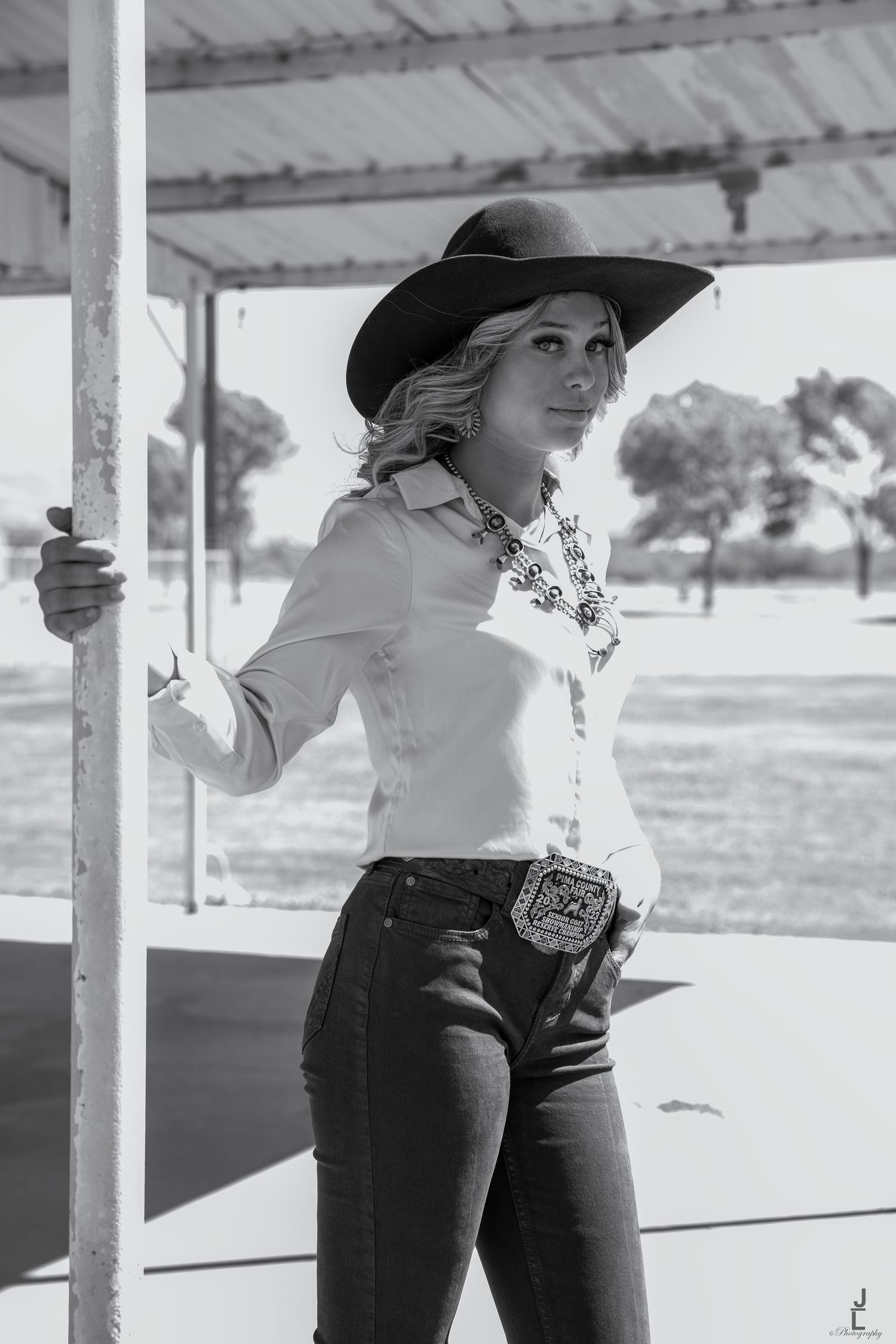 Black and white photo of blonde woman in white shirt and black cowboy hat holding a post outdoors.