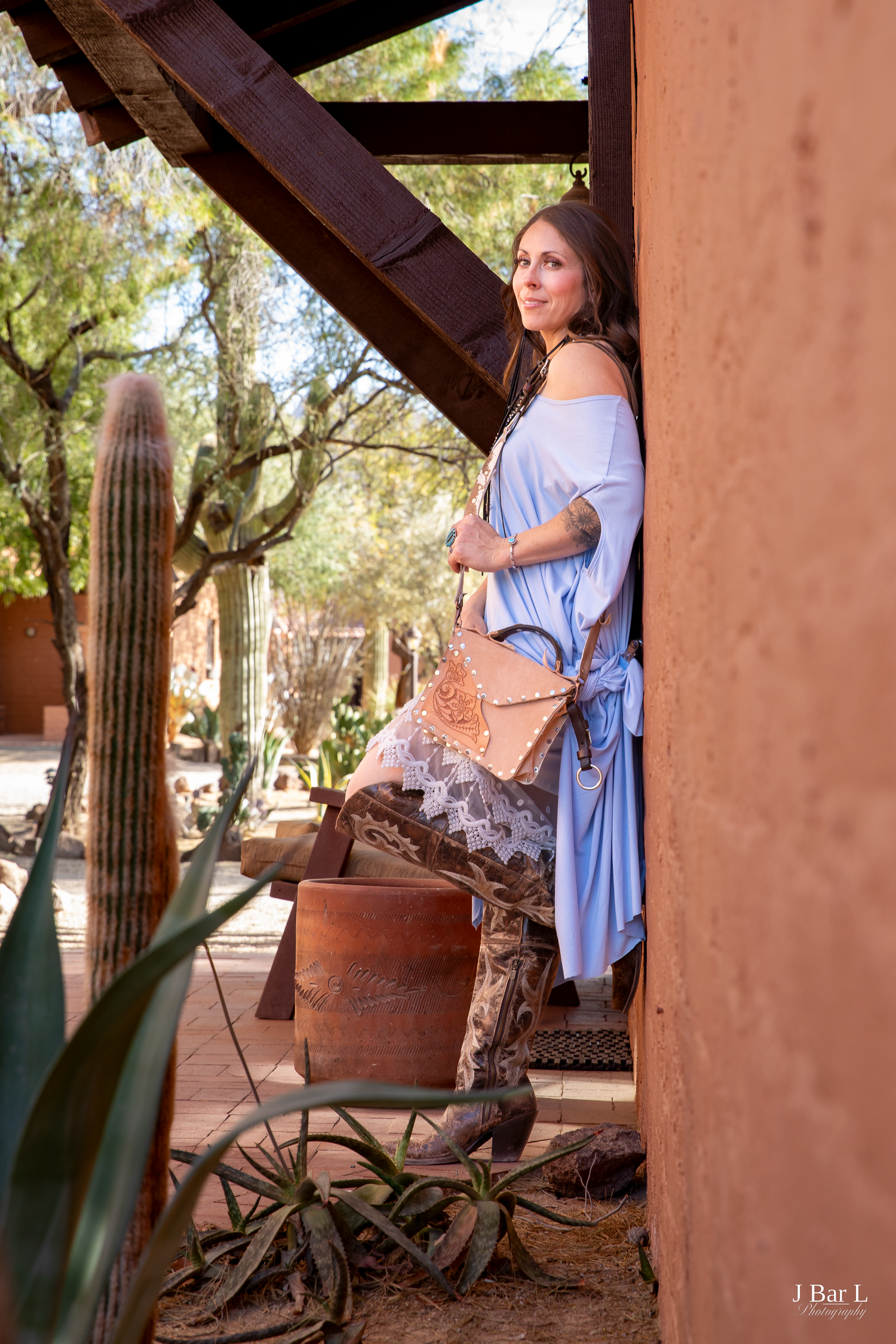Woman in blue dress with tooled leather bag leans against adobe wall near saguaro cactus in desert Southwest.