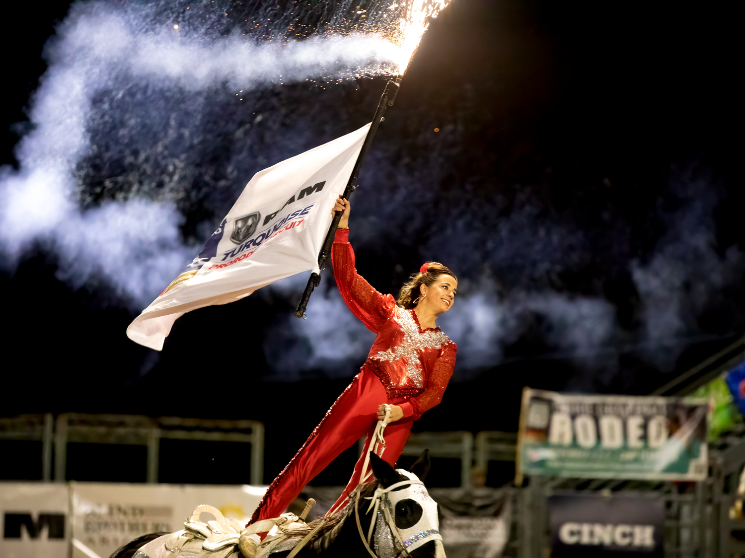 A performer in red holds a flag amid dramatic pyrotechnic explosions during a thrilling nighttime rodeo show.