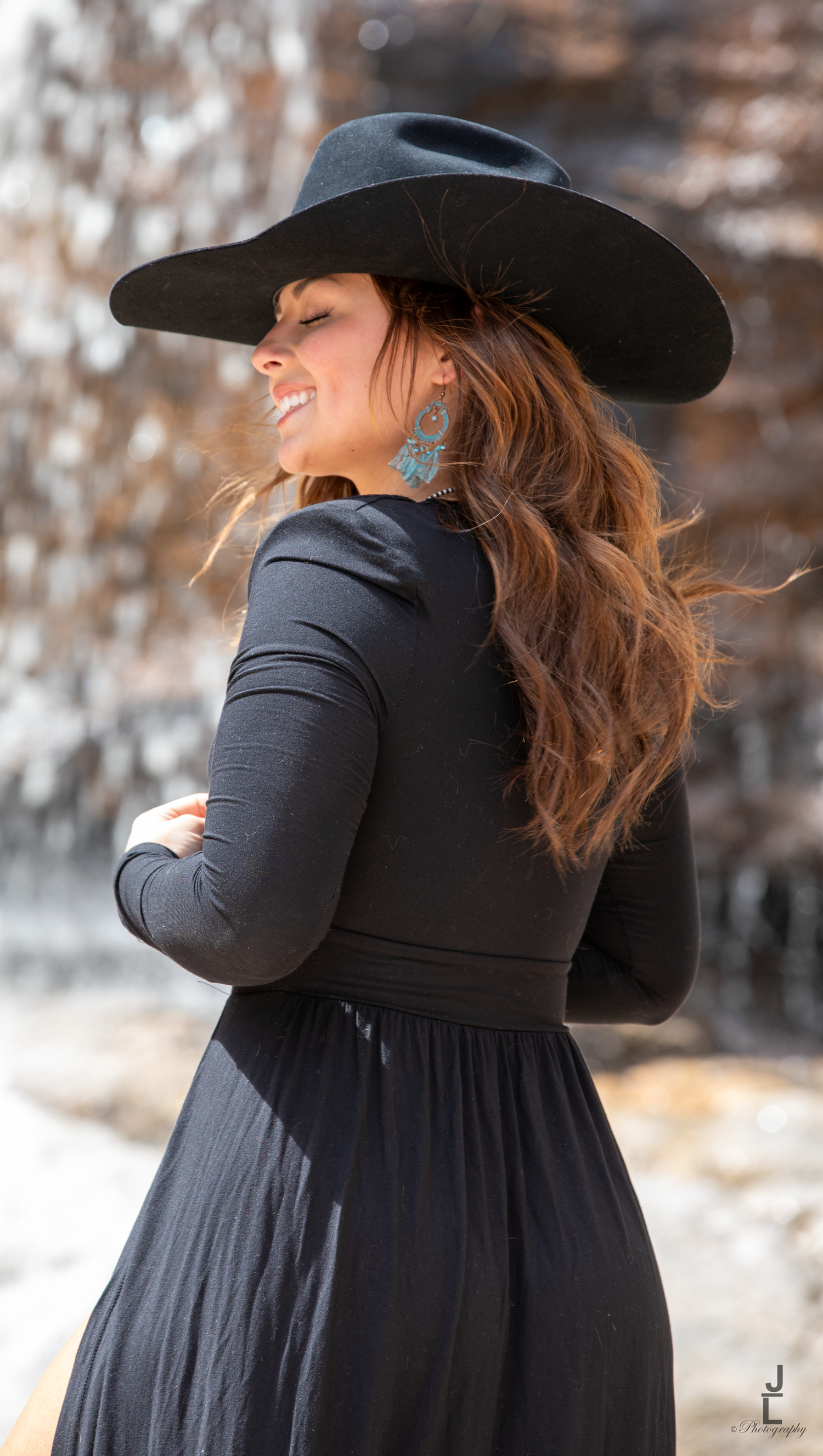 Smiling woman in black wide-brim hat and dress with turquoise earrings outdoors.