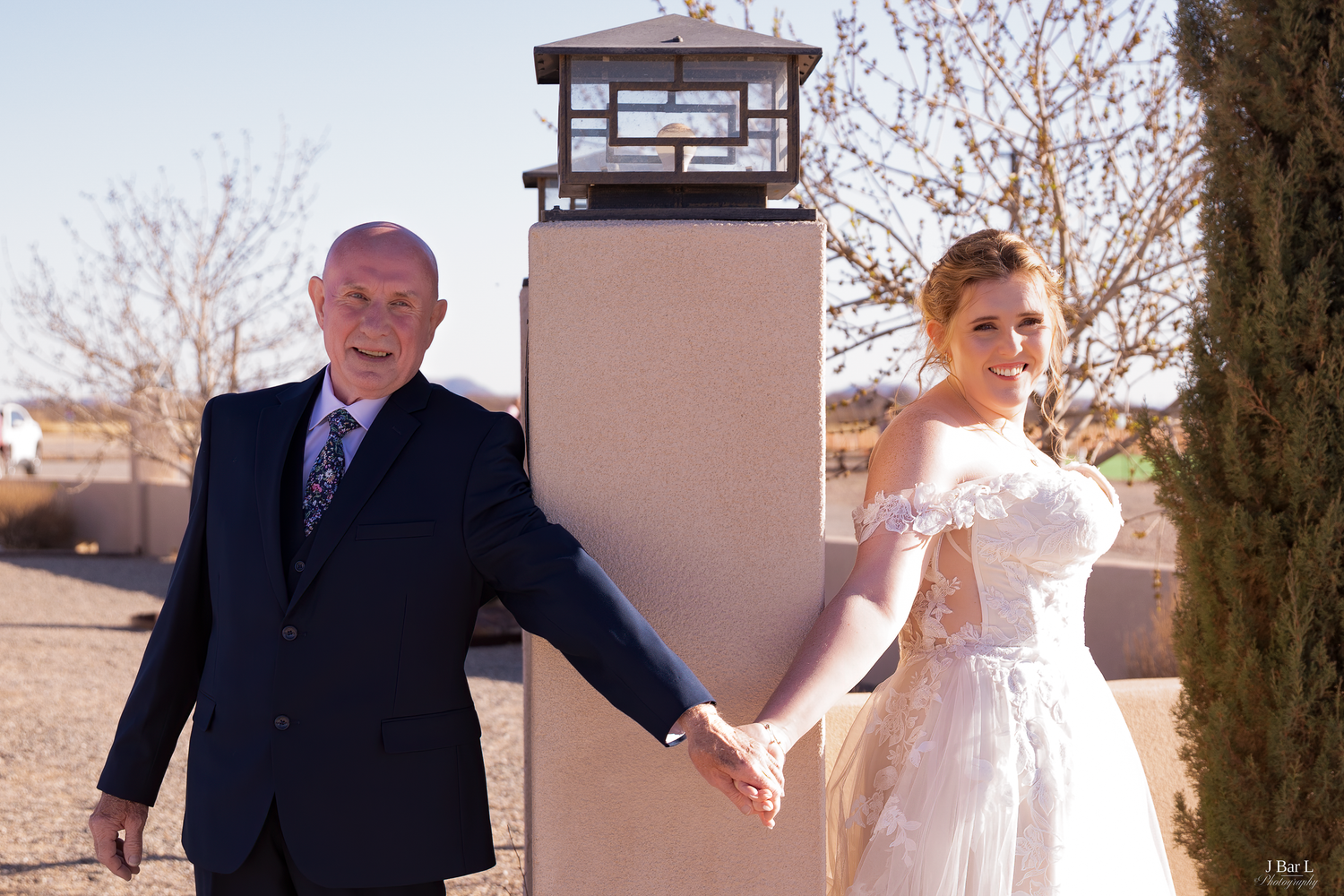 Bride in lace gown holding hands with older man in navy suit at outdoor venue.