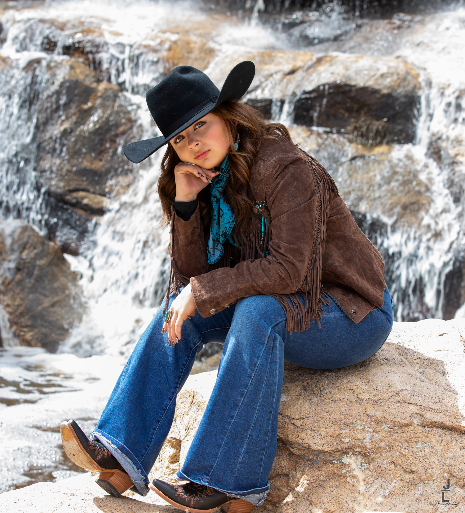 Woman in cowboy hat, fringe jacket, and flare jeans sitting on rocks near a waterfall.