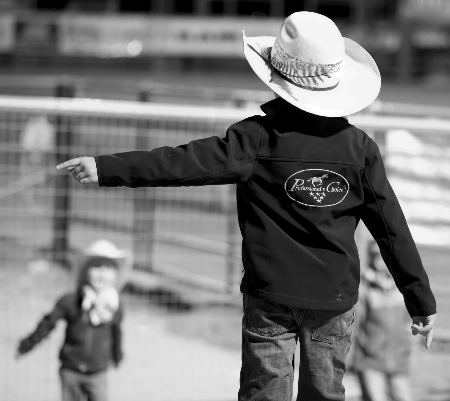 A young child in a cowboy hat and branded jacket gestures at a rodeo event in a black and white photo.
