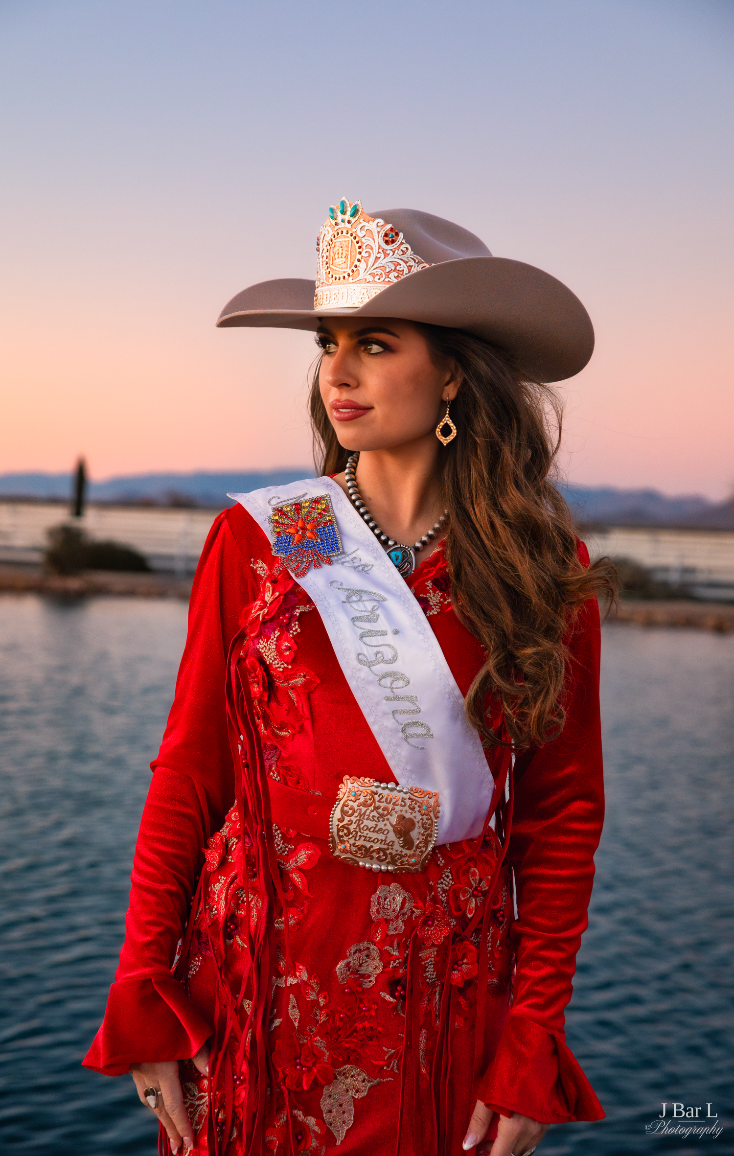 Rodeo queen in a red embroidered outfit with sash and jeweled cowboy hat posing at sunset.