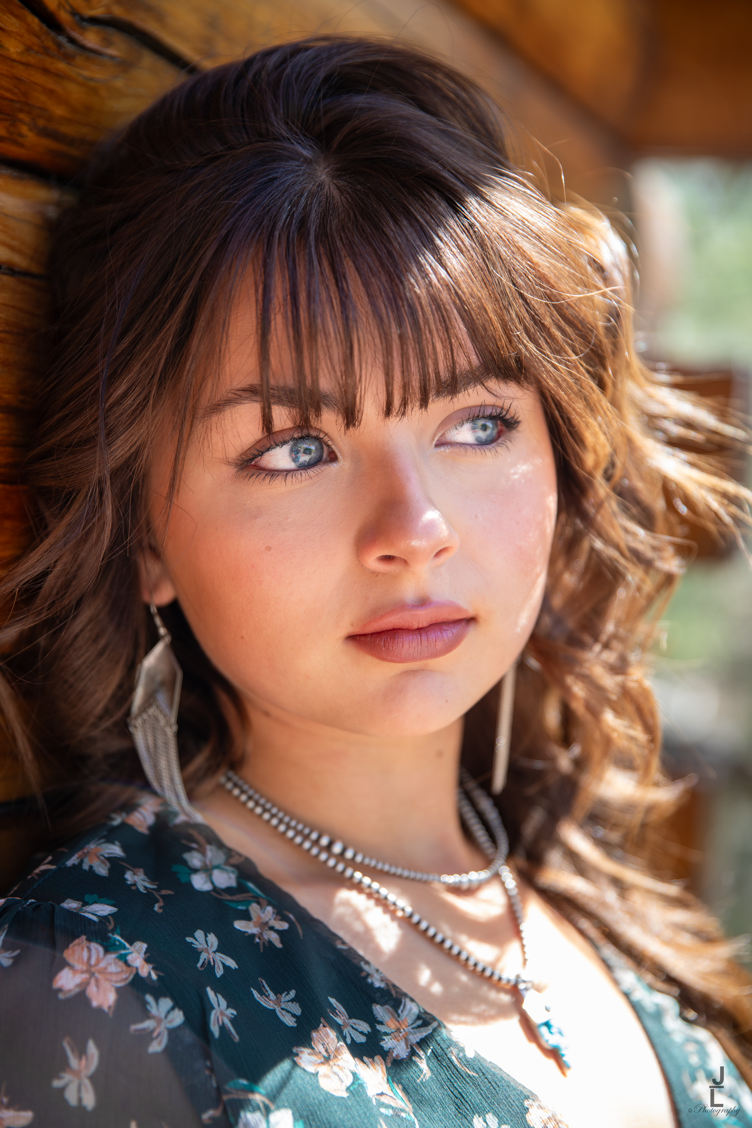 Close-up portrait of a young brunette woman with blue eyes wearing a floral dress and layered necklaces.