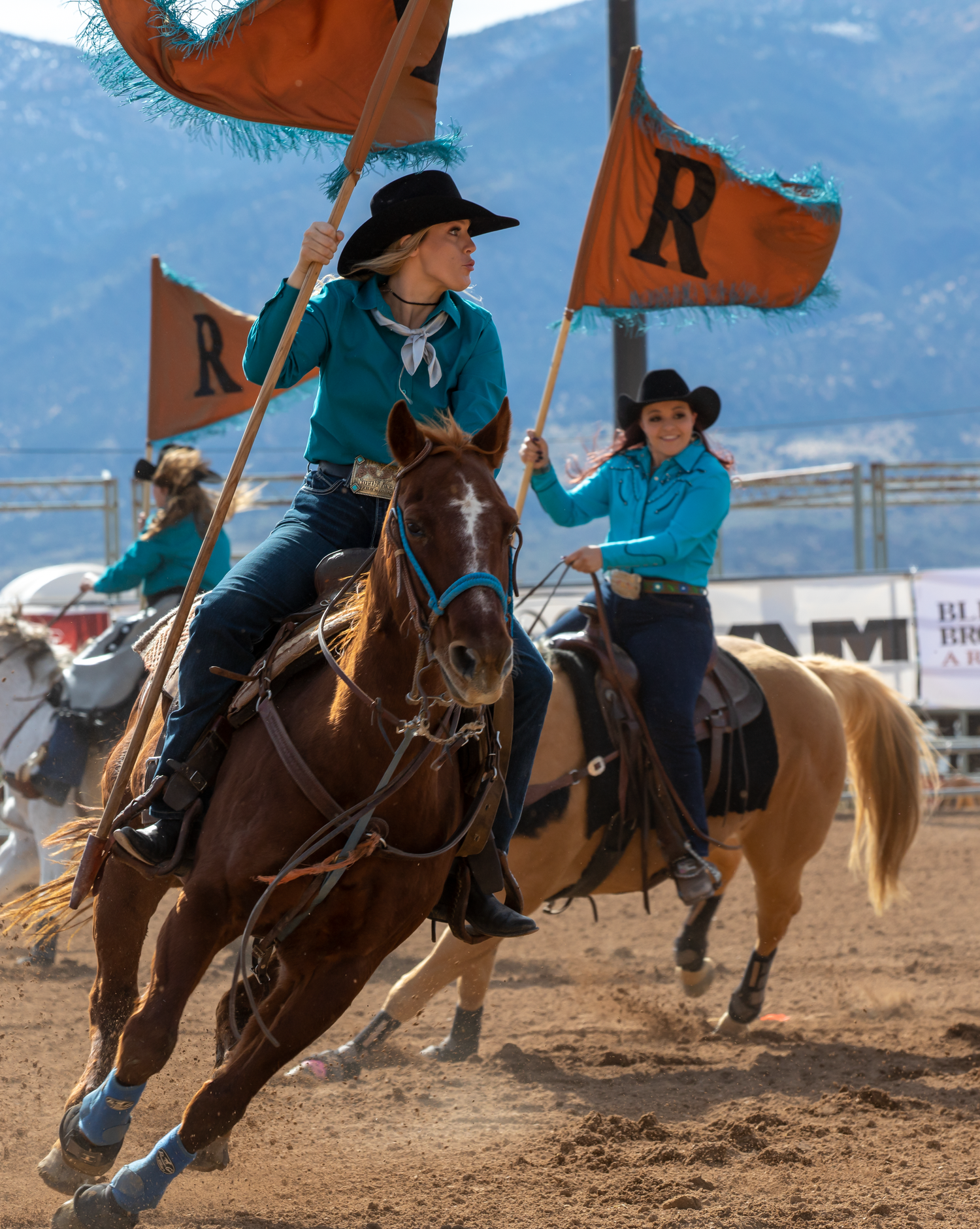 Two female riders in teal shirts carry orange R flags on horseback at a rodeo opening ceremony.
