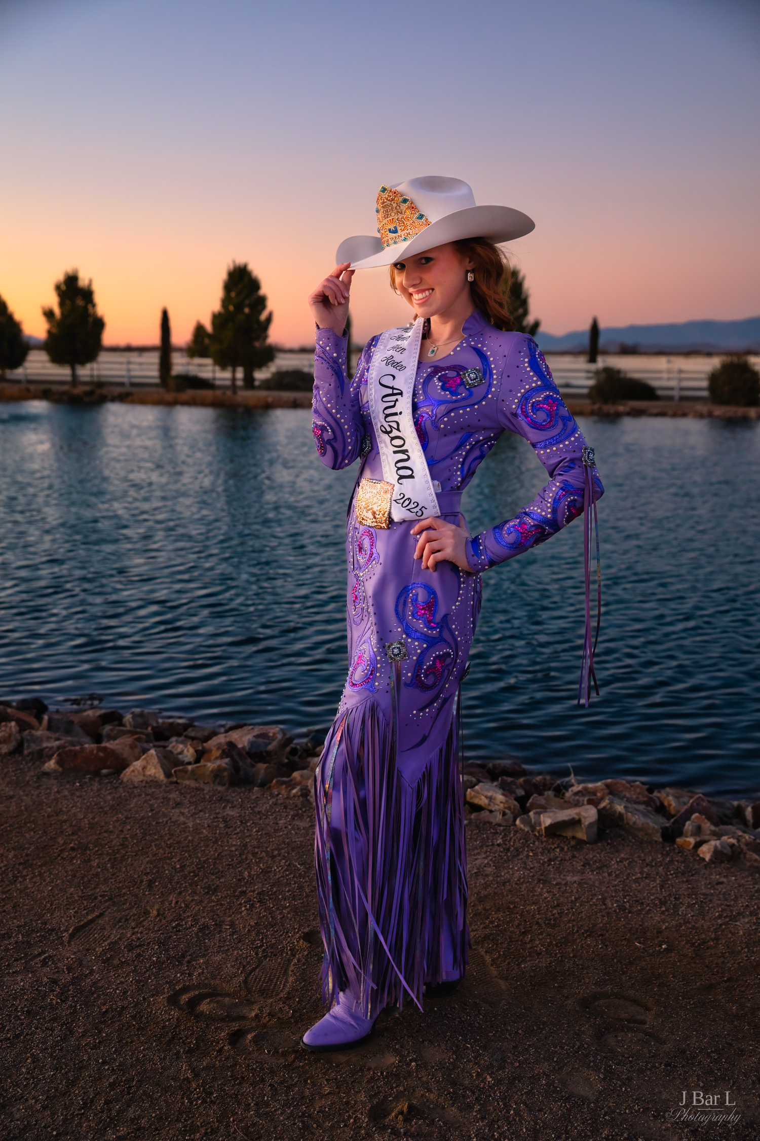 Miss Arizona rodeo queen in purple fringe outfit and white cowboy hat poses at sunset lakeside.