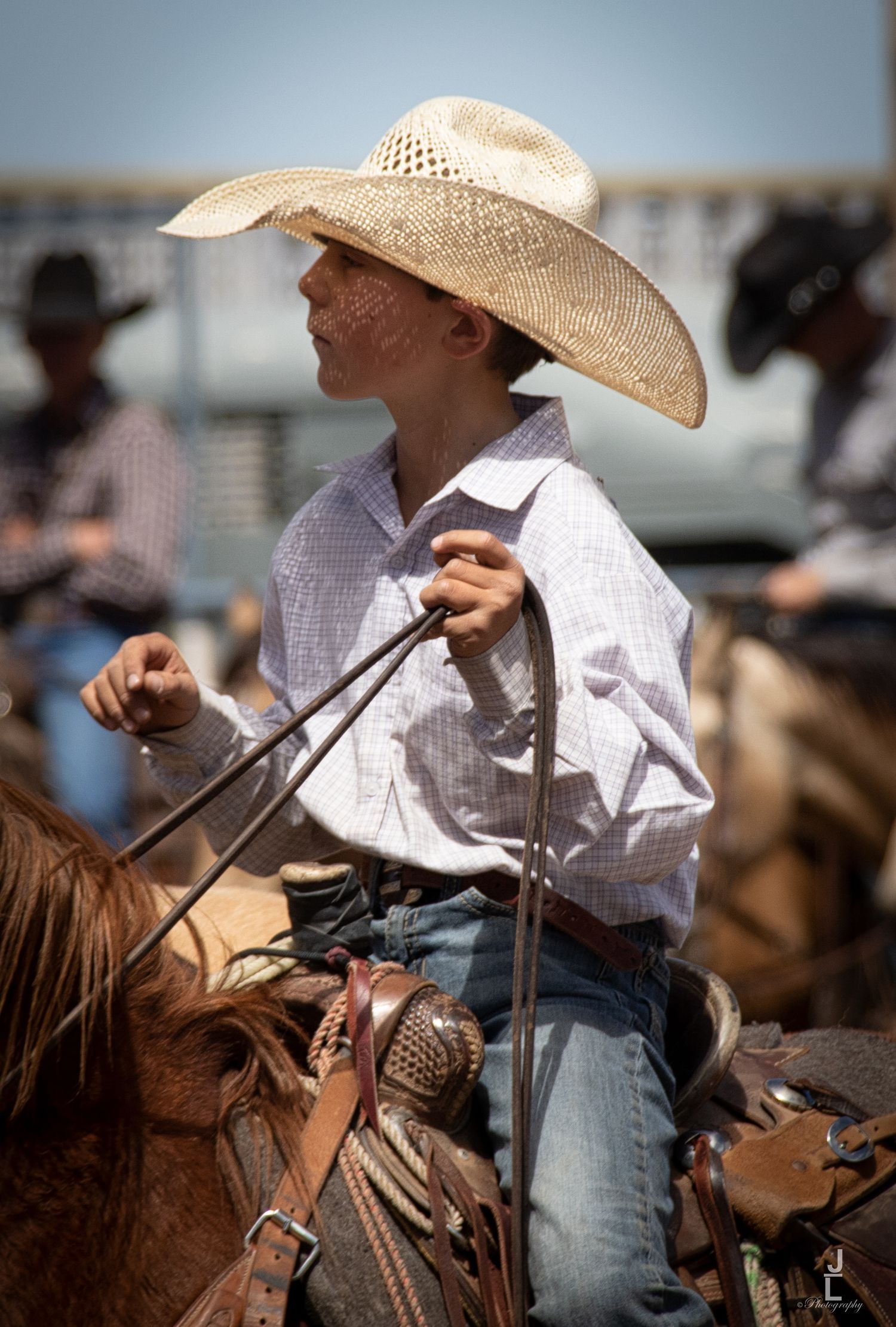 Young cowboy in straw hat sits atop a horse holding reins at a sunny outdoor rodeo.