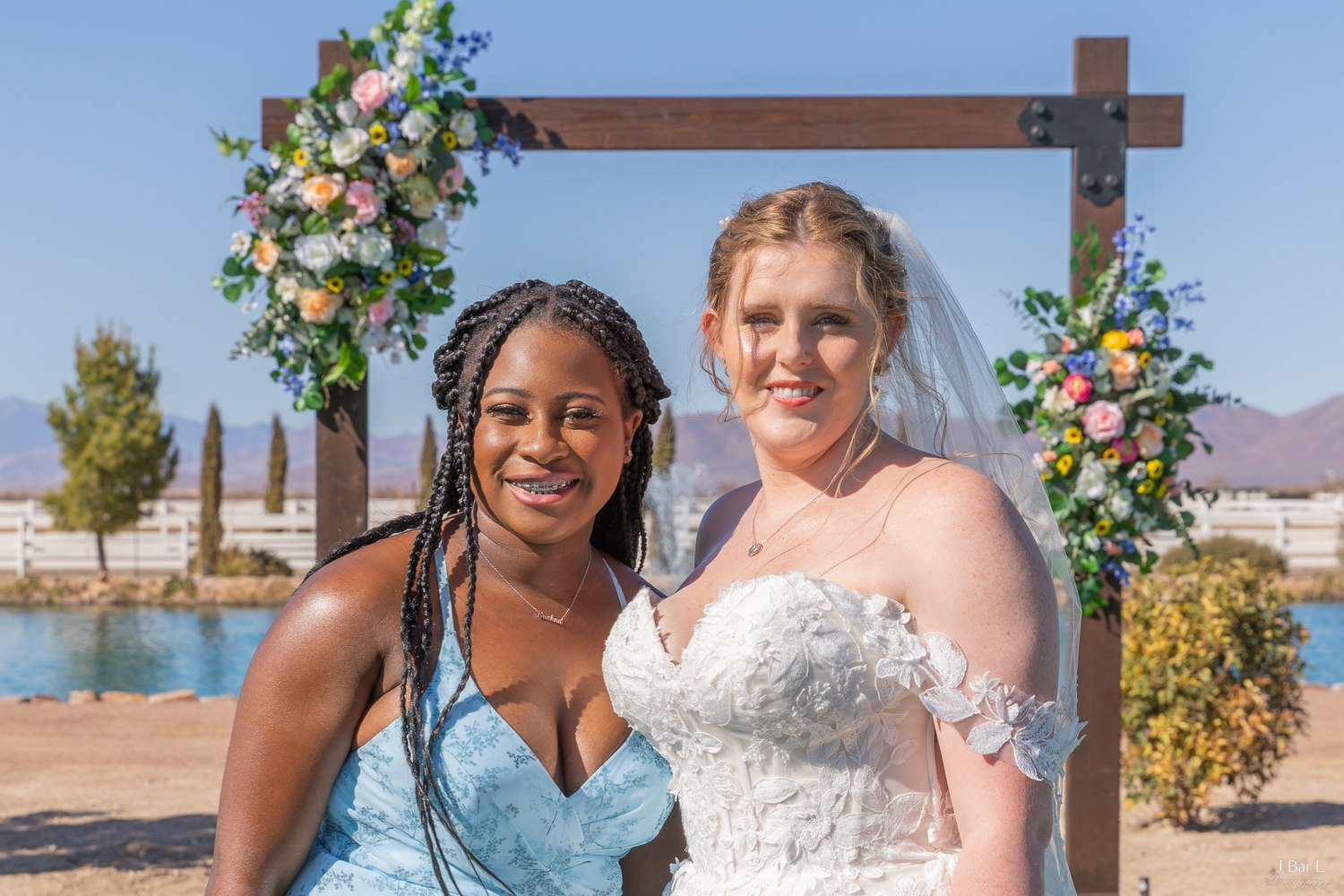 Two women smiling together at outdoor wedding ceremony under floral arch.