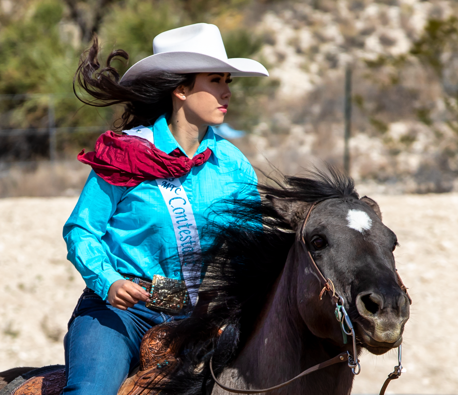 Female rodeo queen in turquoise shirt and white cowboy hat rides dark horse at full gallop.