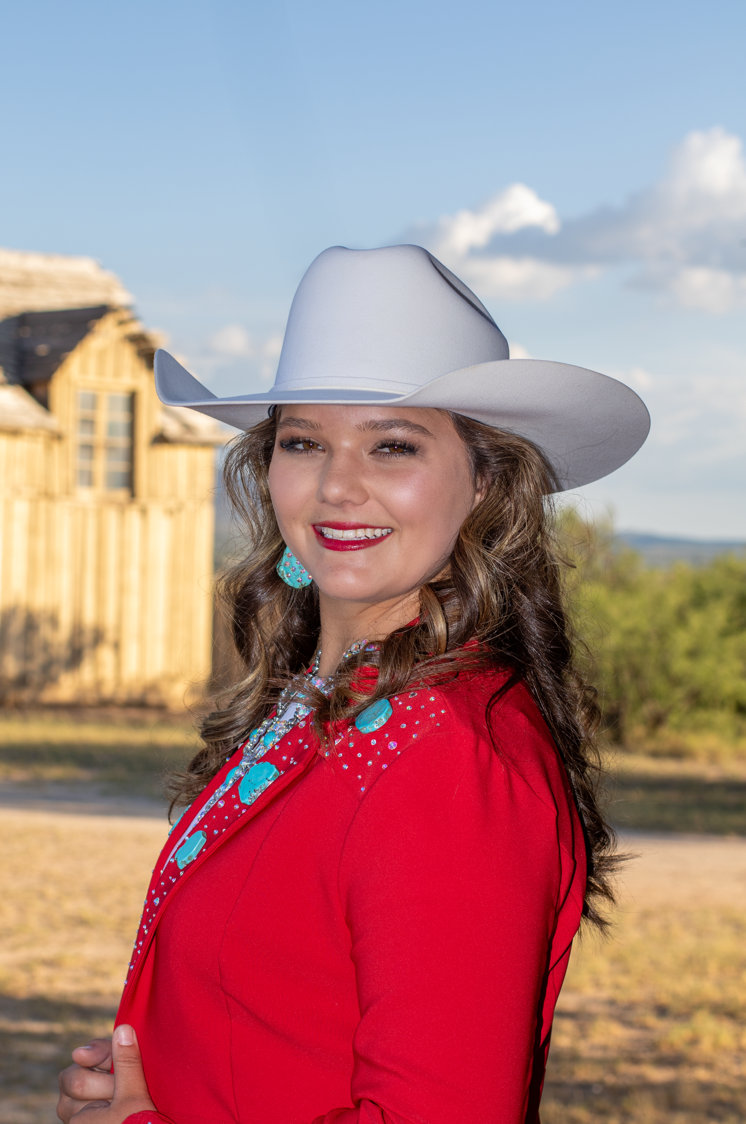 Smiling woman in a red jacket and white cowboy hat with turquoise jewelry at a rustic outdoor venue.