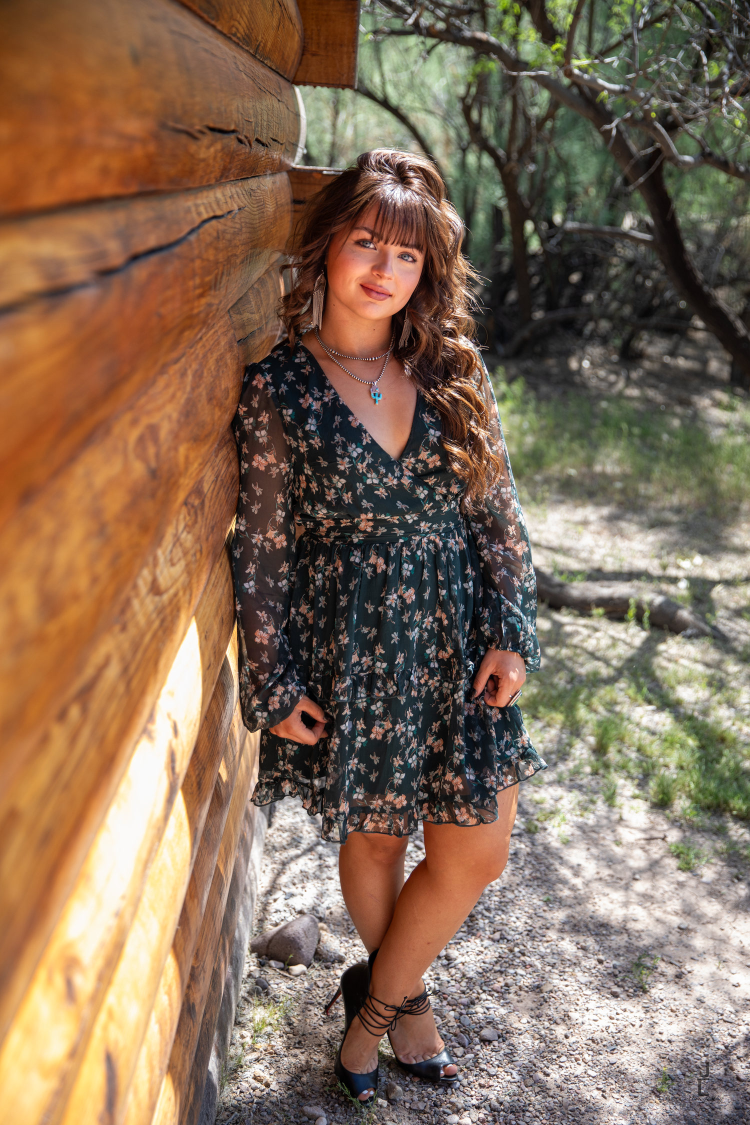 Young woman in floral dress leans against rustic log cabin wall surrounded by desert woodland.